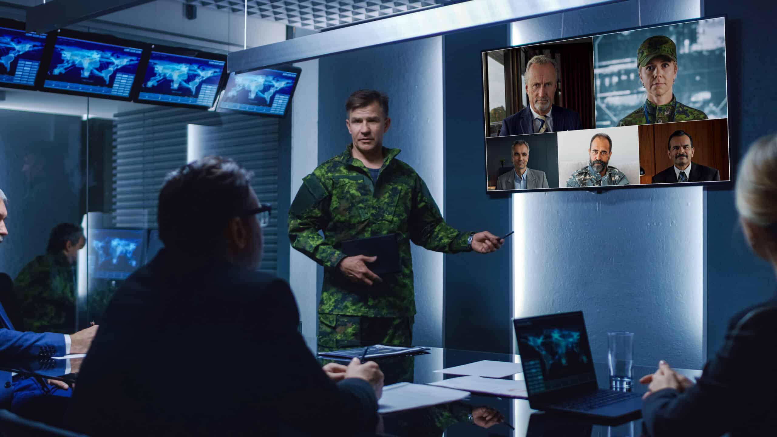 A military person points to a screen showing a video call with four people while three in business attire watch attentively.