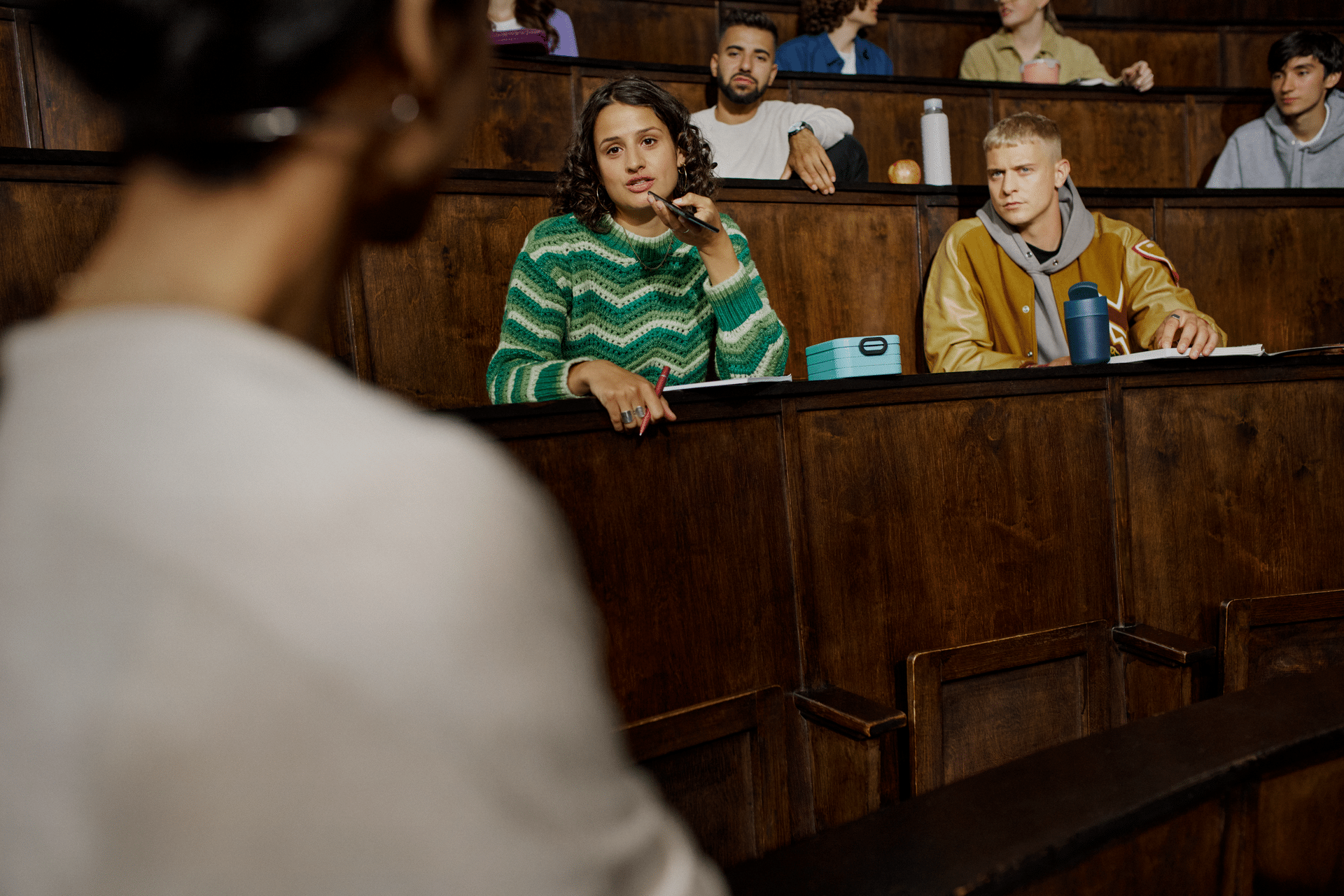 A student in a green sweater raises her hand in a lecture hall, engaging with the instructor as peers listen attentively.