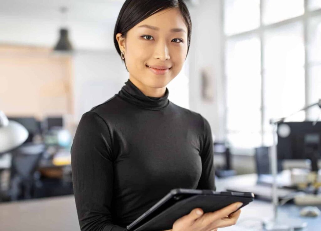 A person in a black turtleneck holds a tablet, smiling in a bright office with large windows behind them.