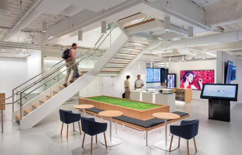 Modern office: glass staircase, green table, screens. Two converse by counter; one ascends stairs. Blue chairs encircle front table.