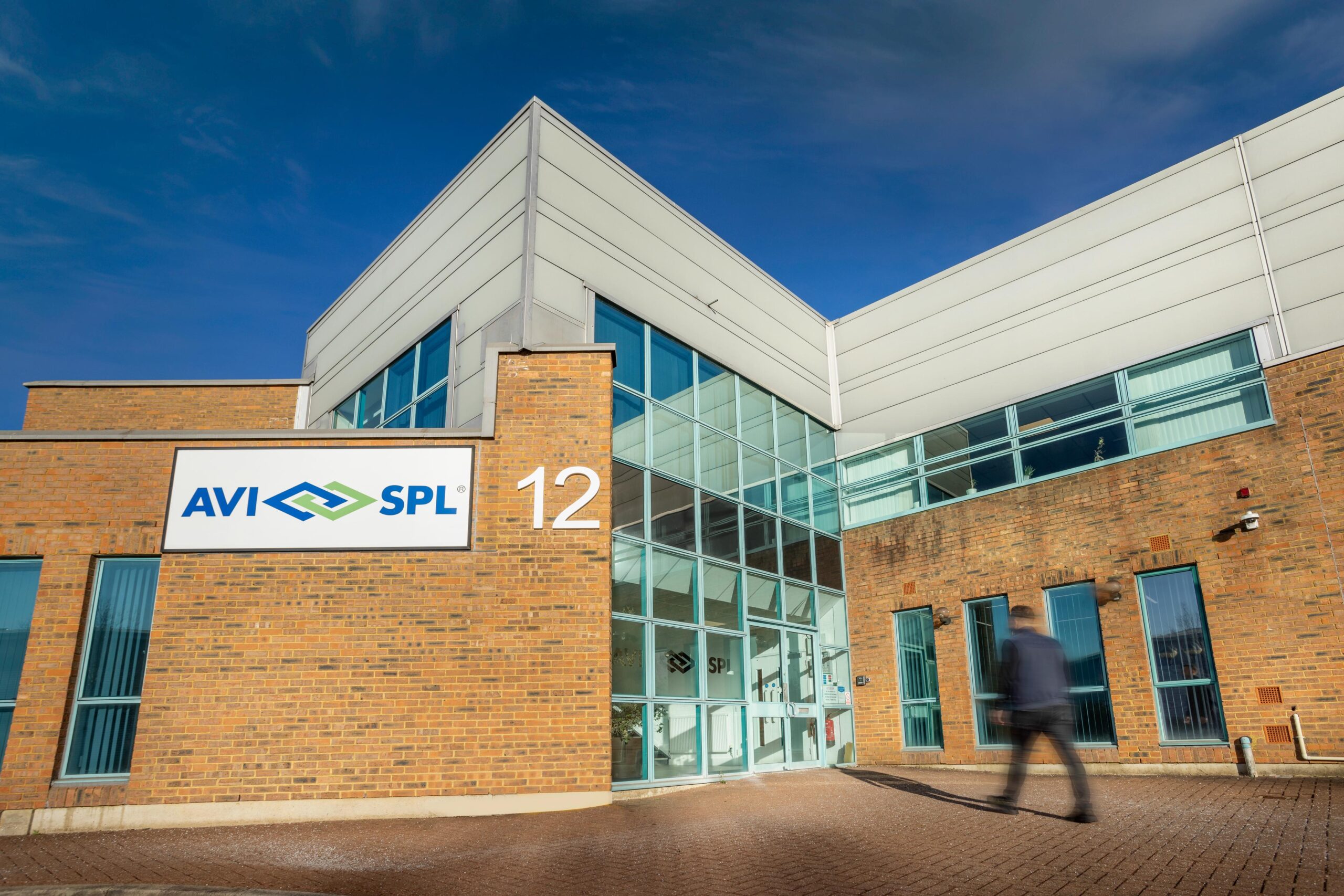 A modern brick and glass building displays the AVI SPL logo and the number 12 as a person walks by under a clear blue sky.