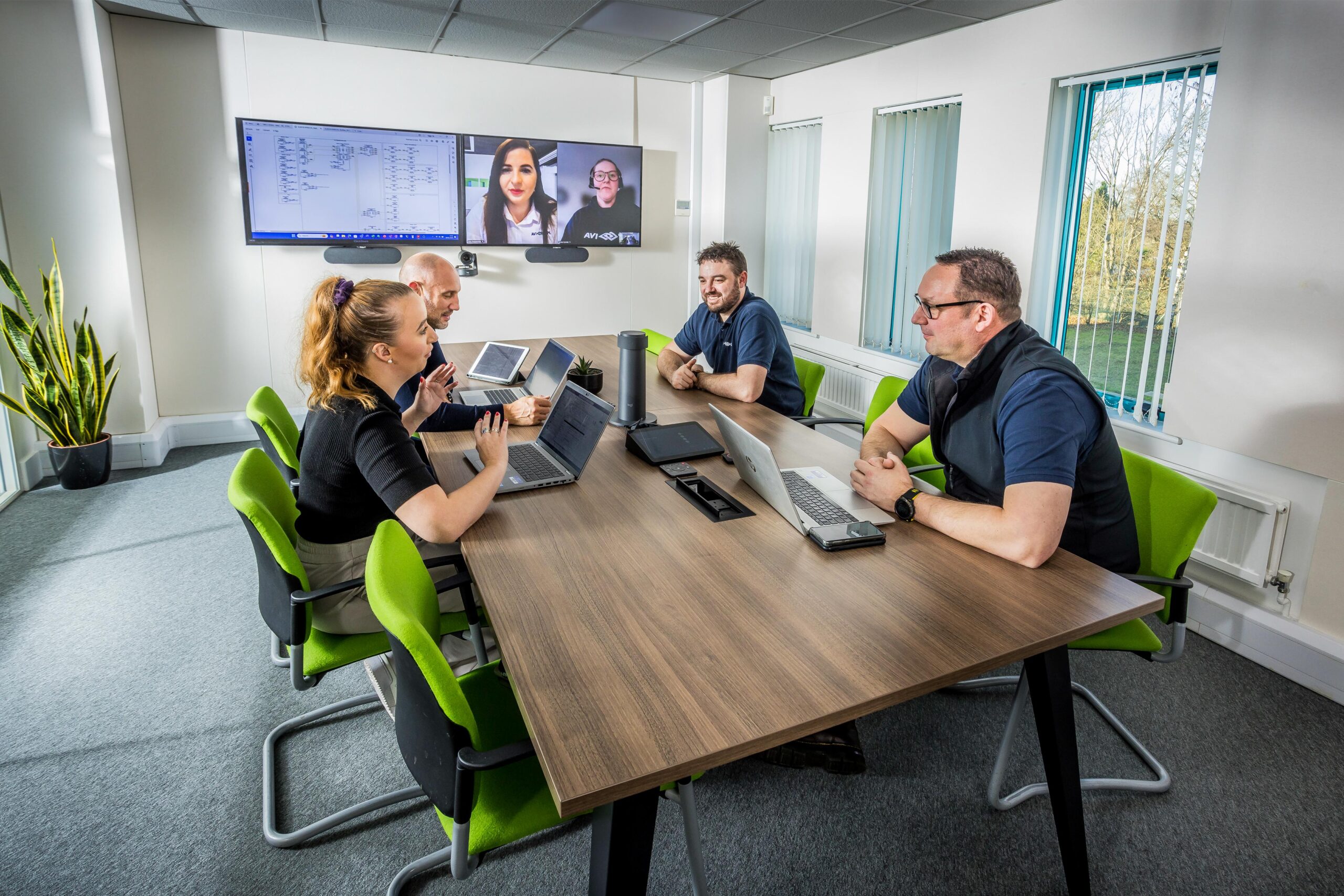 Four colleagues in a conference room on a video call, laptops open. A plant and windows enhance the collaborative atmosphere.