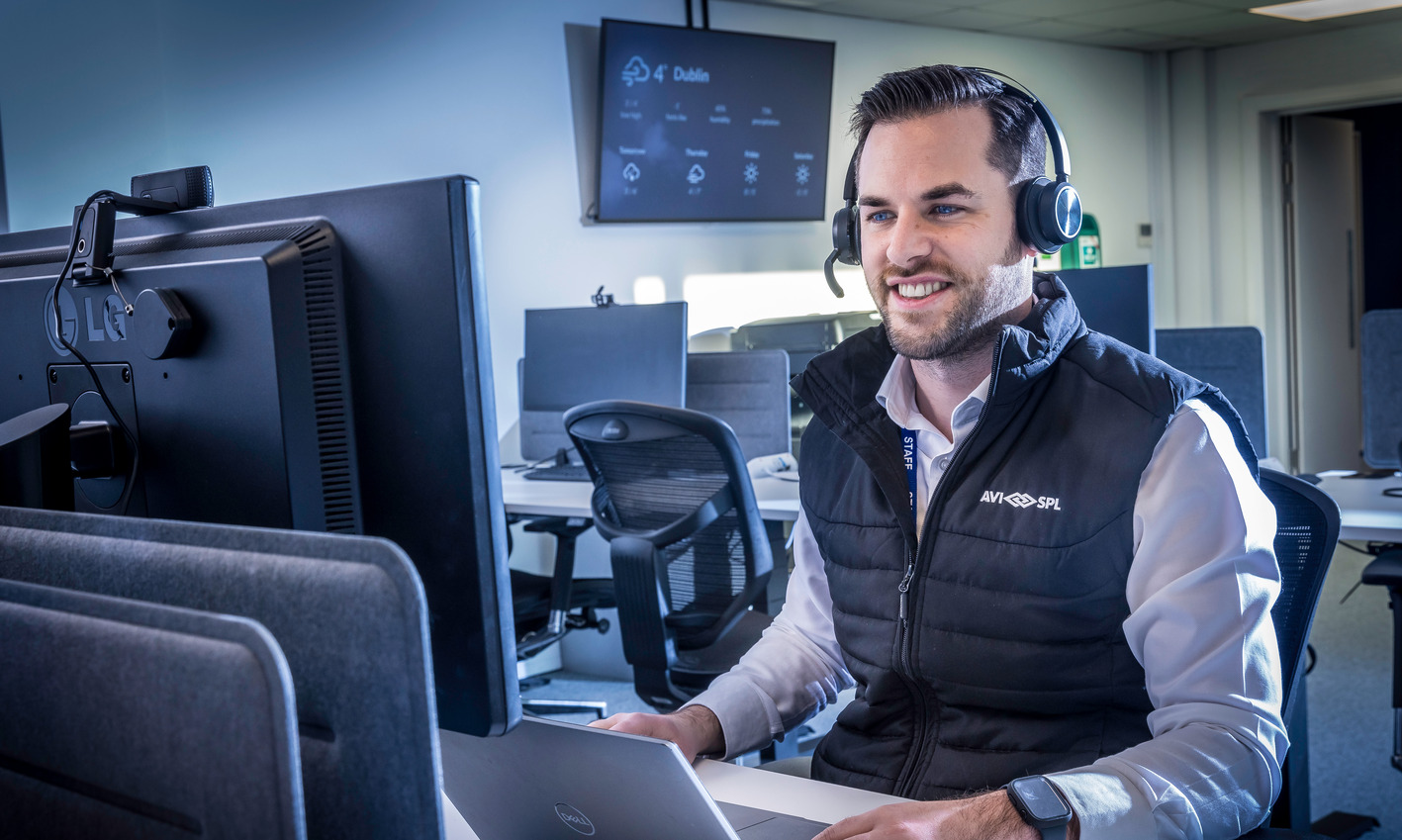 A man in a black vest and white shirt smiles at a computer, wearing headphones at an office desk; empty desks behind.