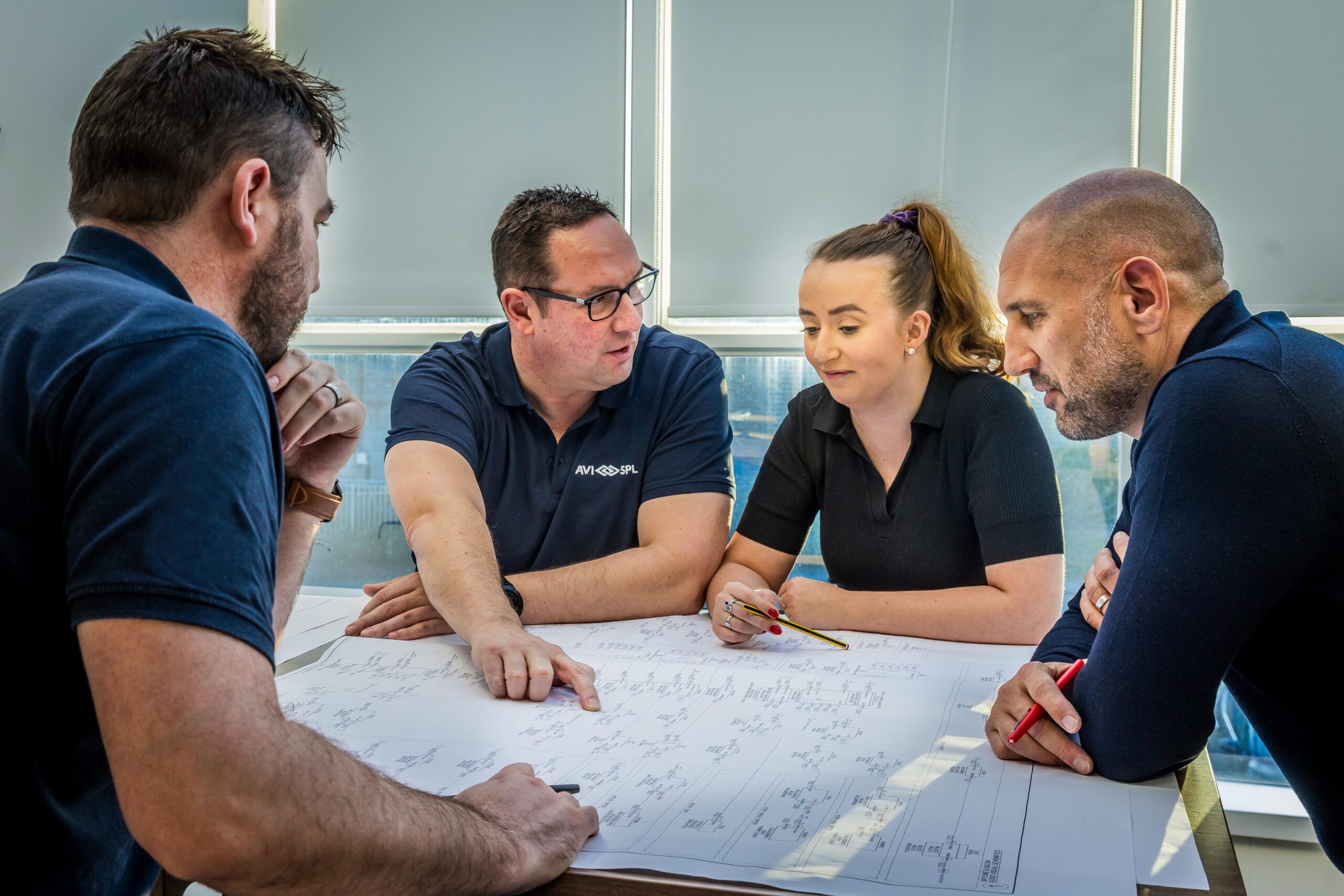 Four people study blueprints; one points to a section. Others watch intently in a well-lit room with window blinds.