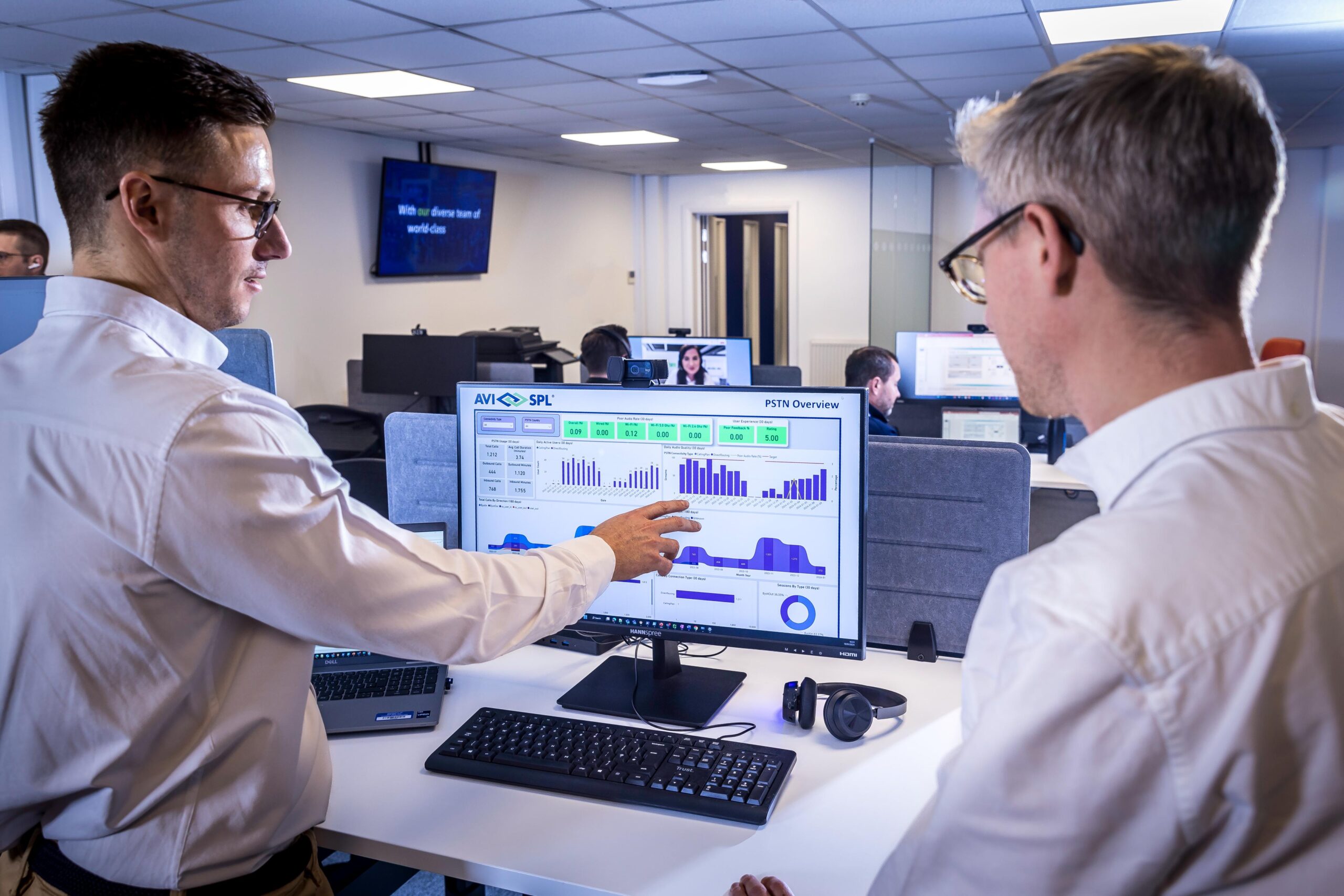 Two men in white shirts analyze data on a computer, pointing at a bar chart. Office background with workstations and wall screen.