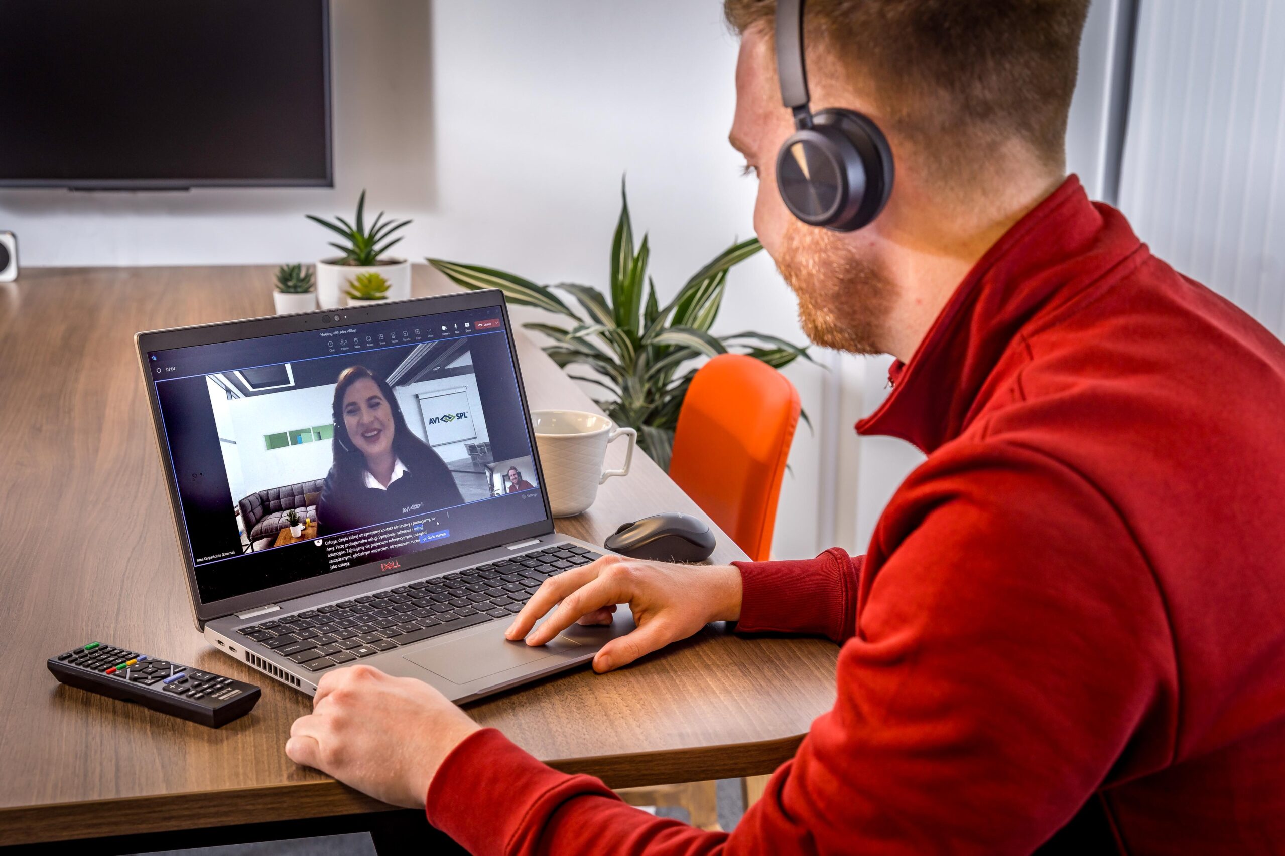 Man on video call with a woman, headphones on, at wooden table with remote and cup; office chairs and plants in background.