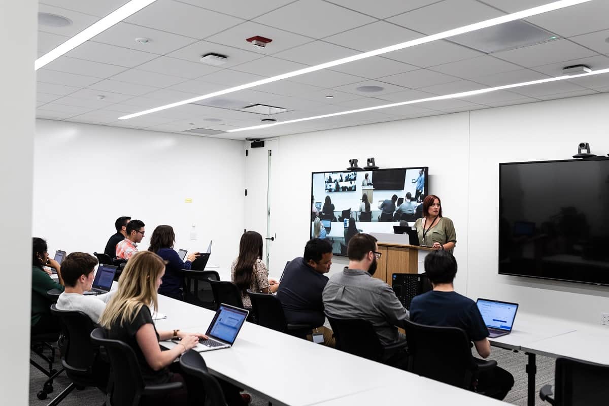 People in a modern classroom, some with laptops. A woman speaks upfront, video call on screen; well-lit with whiteboards and monitors.