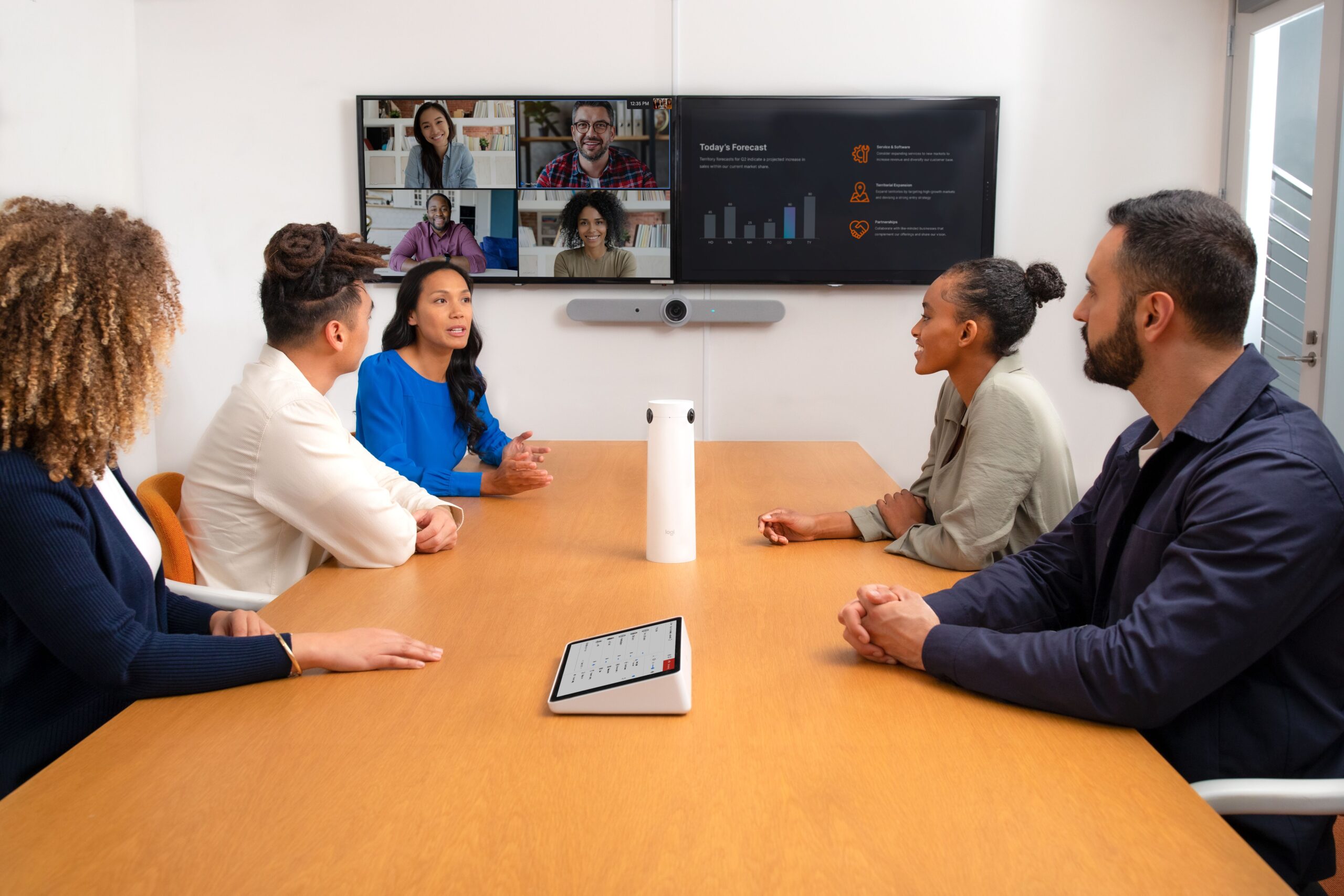 Five people in a modern meeting room are engaged in a video call, with a large screen, tablet, and cylindrical device on the table.