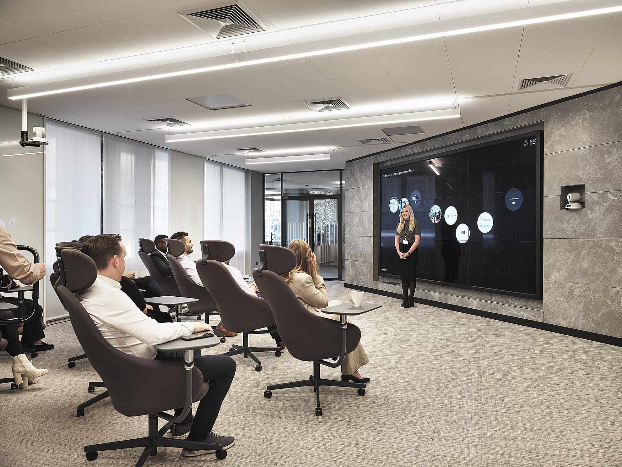 In a modern conference room, attendees sit facing a large screen with circular icons, surrounded by gray walls and windows.