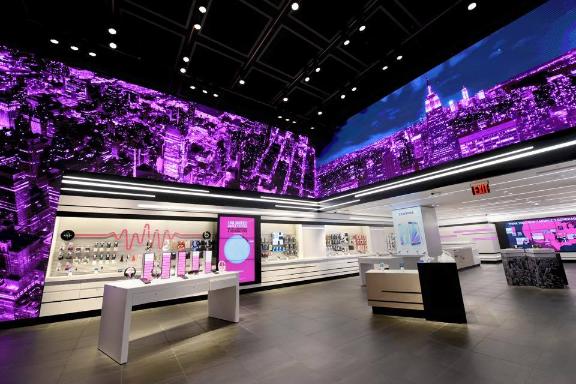 A tech store interior with a large purple cityscape display, gadgets on white shelves, black grid ceiling, and gray tiled floors.