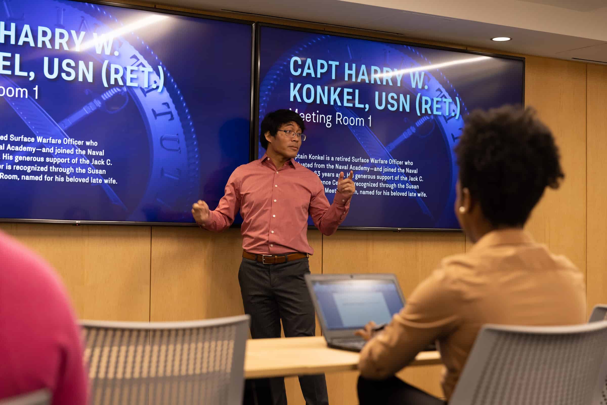 Man in red shirt talks in front of screens; woman with short hair takes notes.