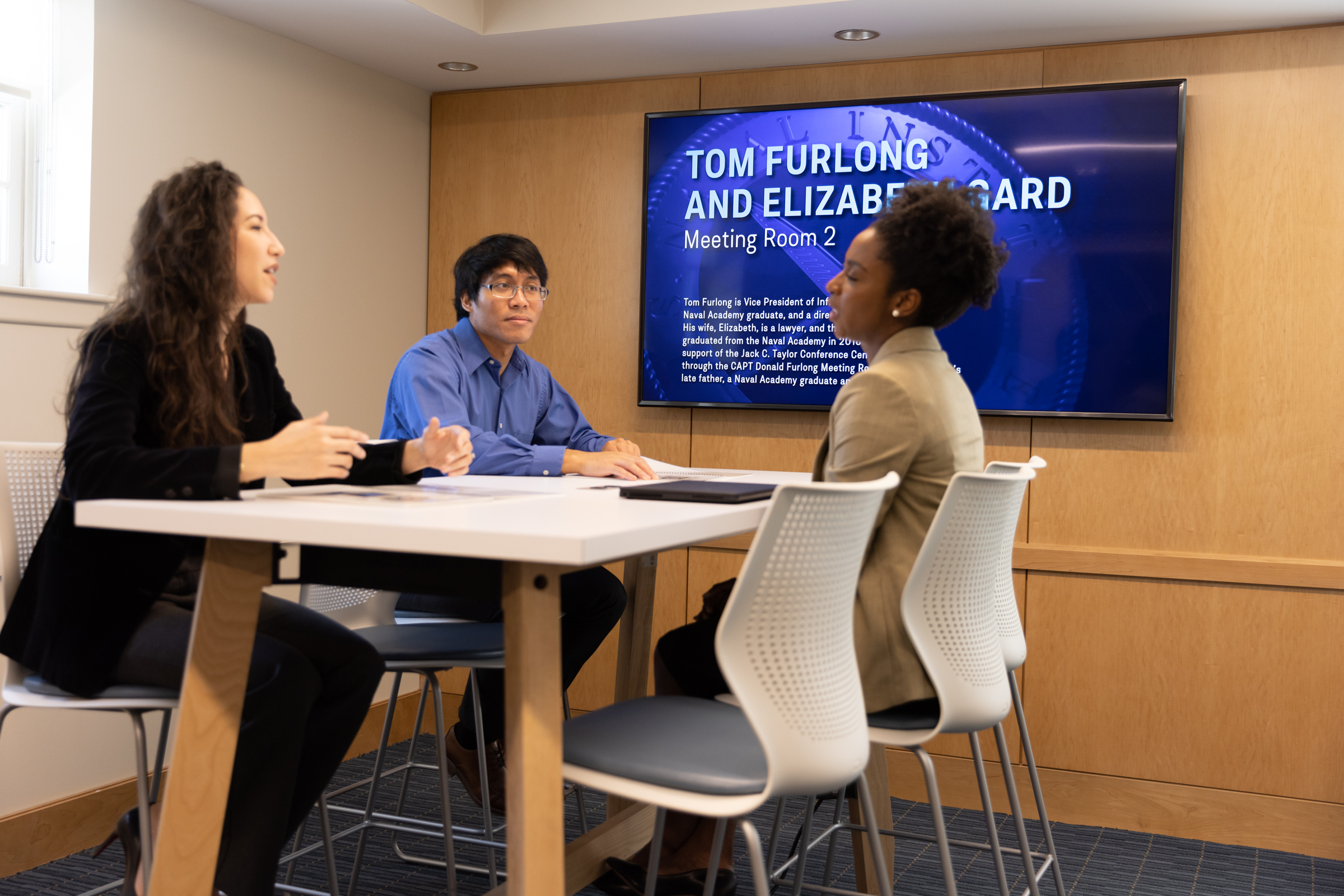 Three people at a table in Meeting Room 2, with focus on a man in blue and woman in dark jacket engaged in discussion.