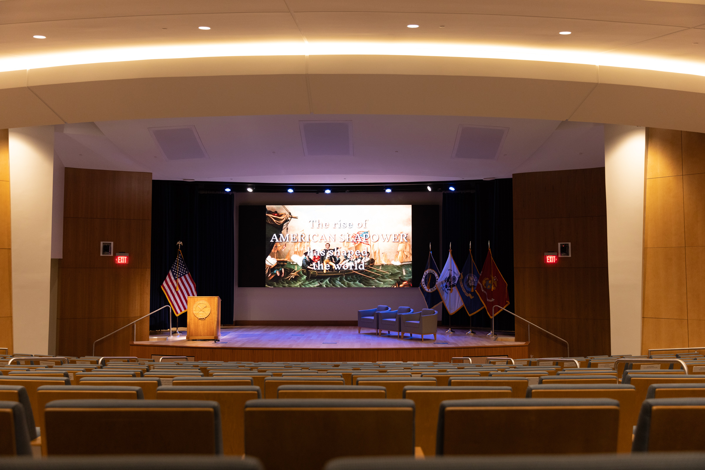 Empty auditorium with rows facing a stage. Screen reads The Rise of American Sea Power. Flags, podium, chairs in front.