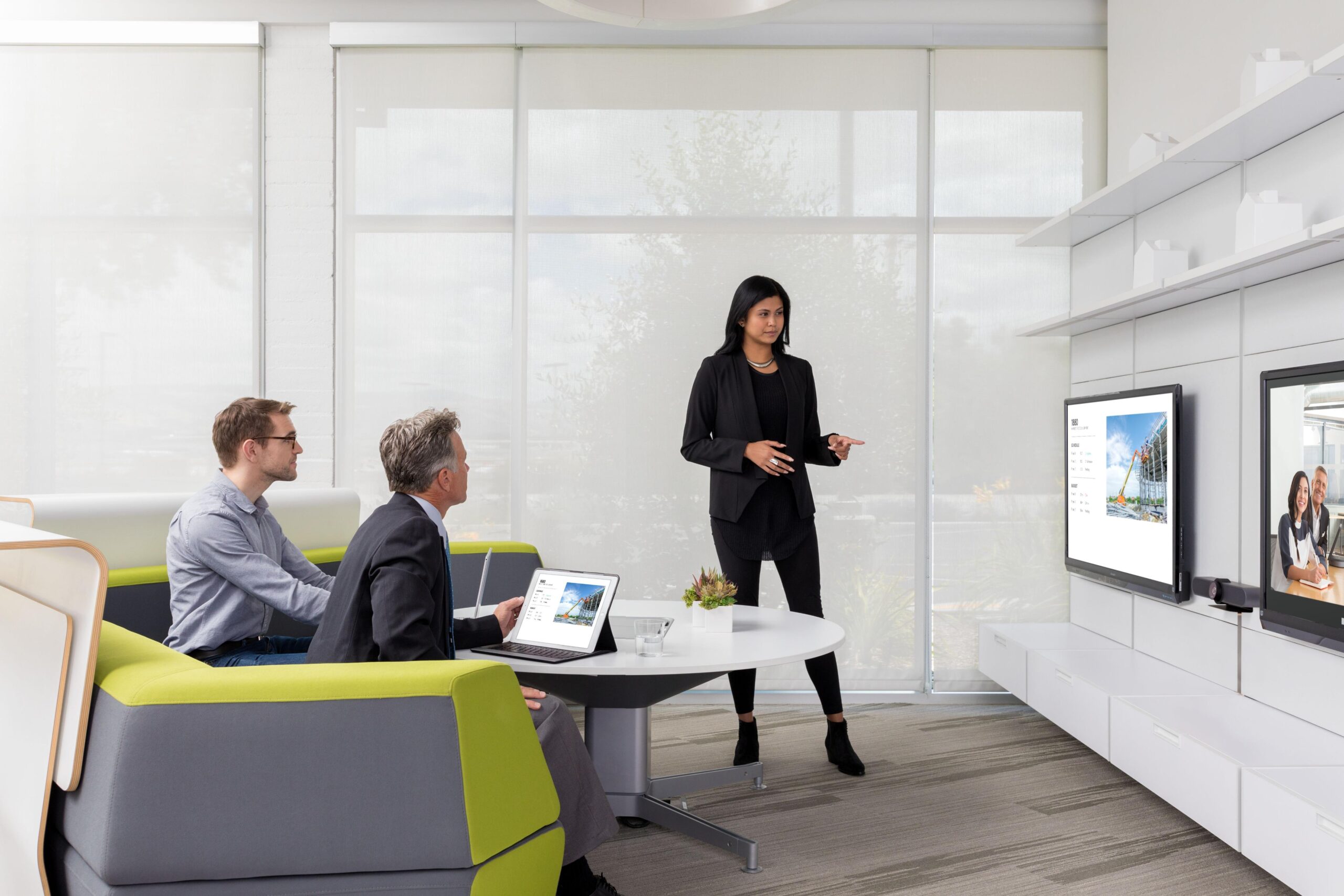 A woman presents to two men in a modern, well-lit meeting room with large windows, using a TV and laptop.