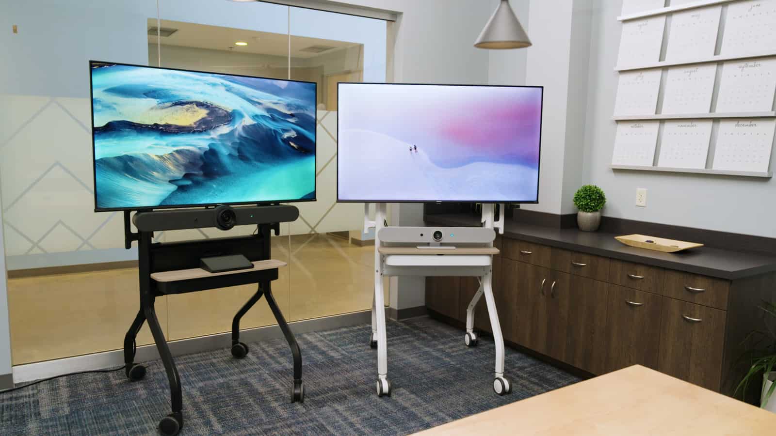 Two digital screens on stands in a modern office, displaying abstract landscapes; features a cabinet, plant, calendar, and glass partition.