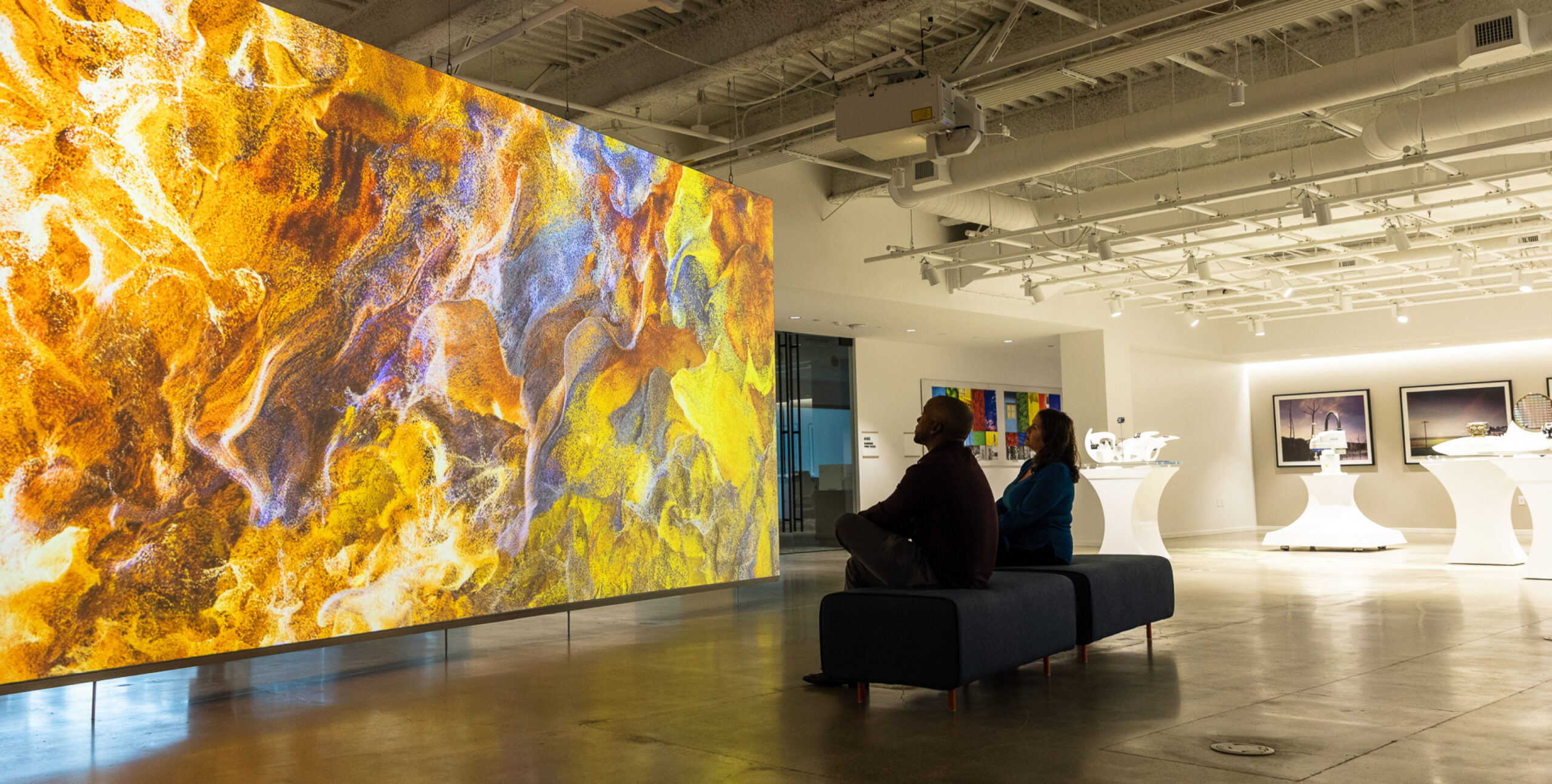 Two sit on a bench in a gallery, facing vibrant abstract art with yellow and purple swirls, surrounded by sculptures.