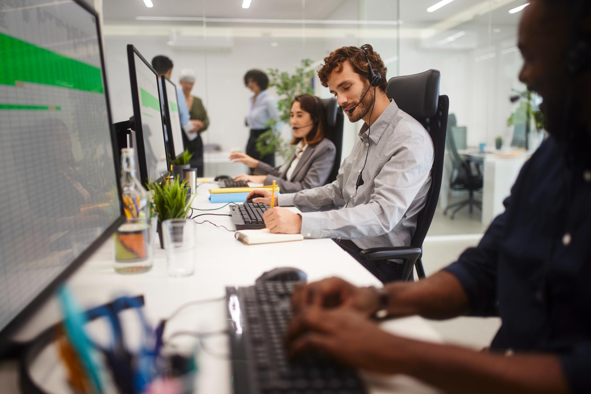 People in an office, wearing headsets, work on computers in a modern, plant-filled space, with more colleagues visible behind.