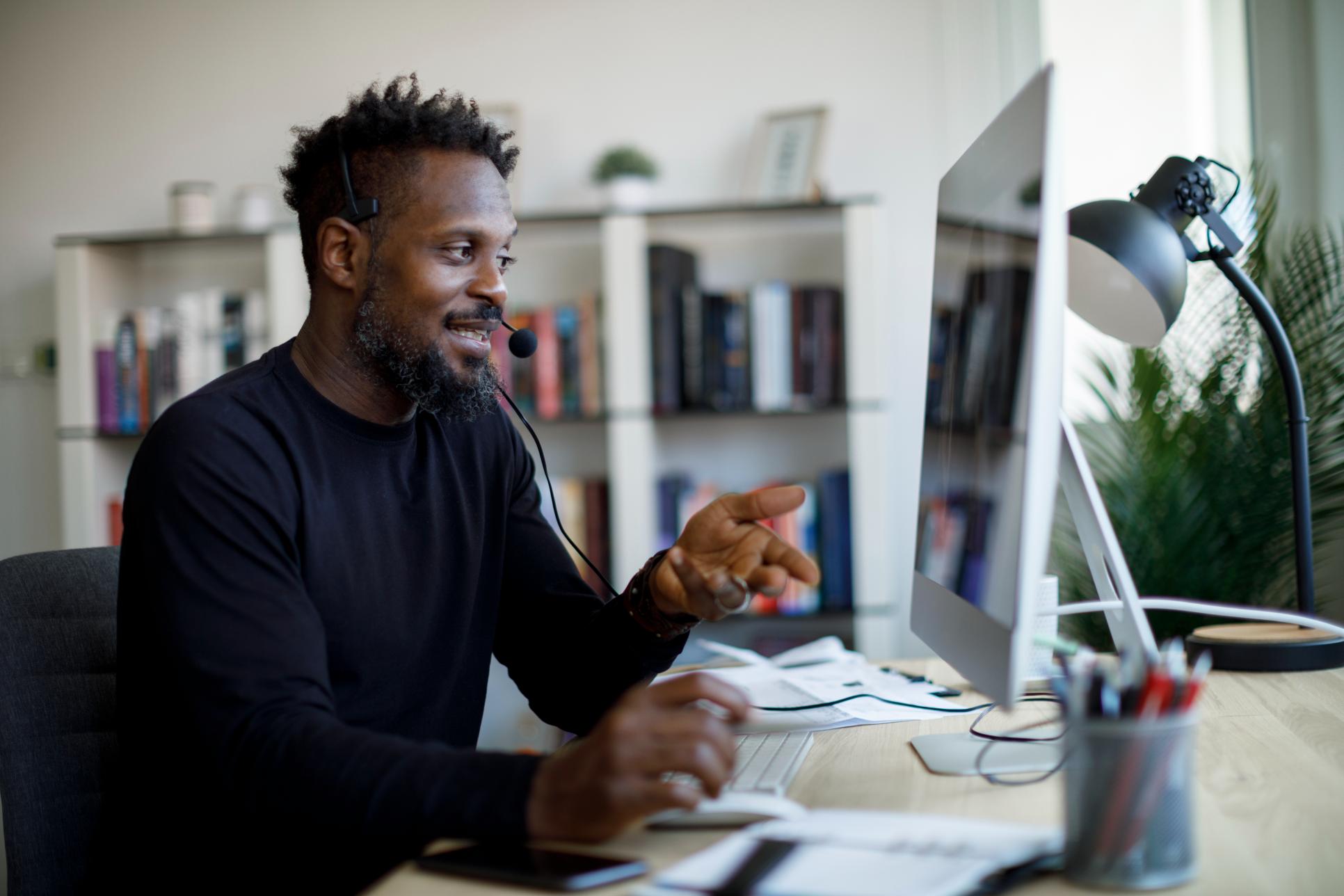 A person in a headset at a desk on a video call, gesturing animatedly. Bookshelves and a lamp in the background complete the scene.