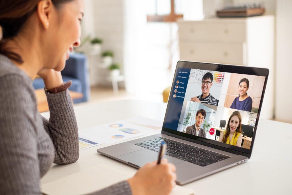 A woman at a desk joins a video call on a laptop with four smiling people, holding a pen and notebook by charts and papers.