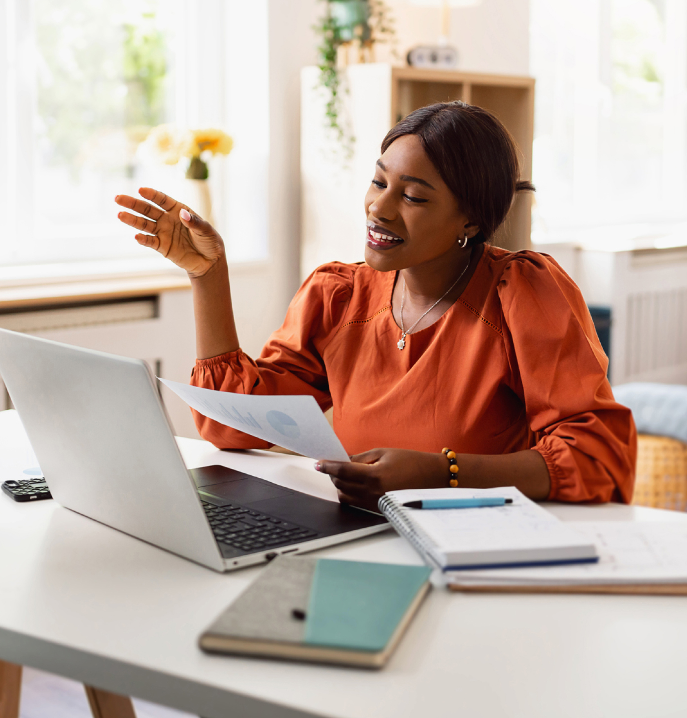 Woman in orange blouse on video call, holding paper. Notebooks and pen on desk; airy room with plants in background.