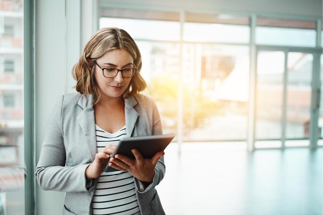 Woman in gray blazer using a tablet in a bright modern office with natural light; short wavy hair, glasses.