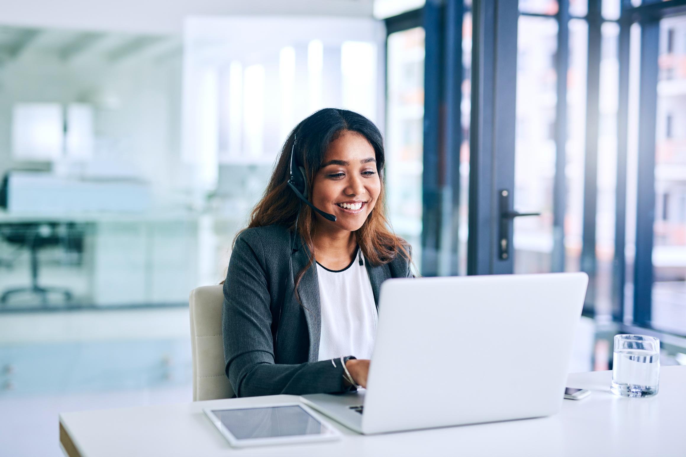 Woman in blazer at desk, smiling with headset and laptop; tablet, water nearby. Bright office with large windows in the background.