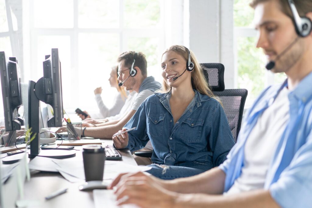 A woman in denim smiles, chatting amidst colleagues wearing headsets, focused on screens in a bright office.