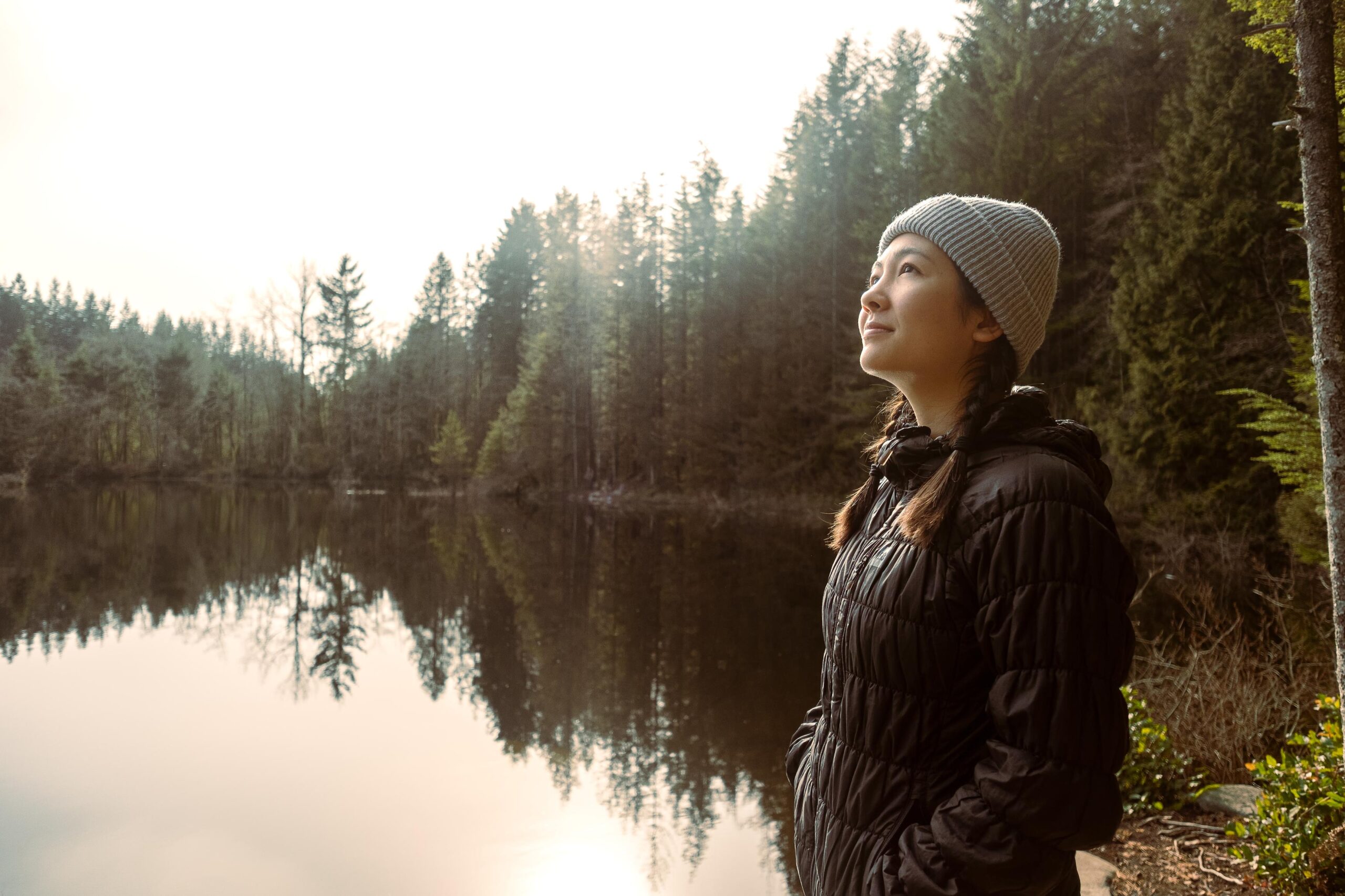 Person in black jacket, gray hat by calm lake with trees. Gazes upward serenely; overcast sky reflects on water.