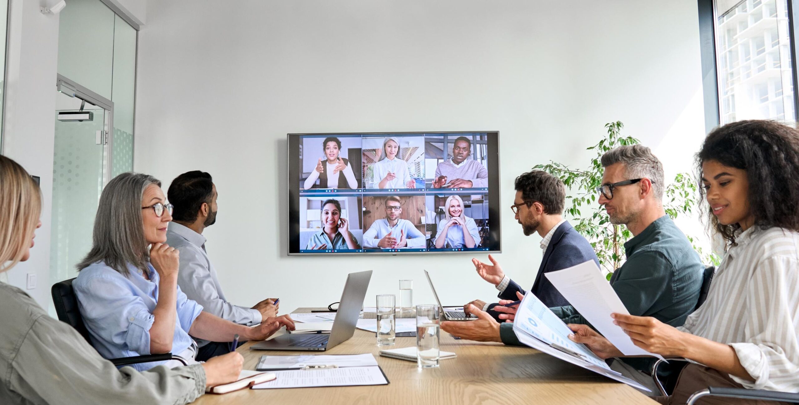 A diverse group of six in a conference room for a virtual meeting, screen with participants; documents, laptops, urban view outside.