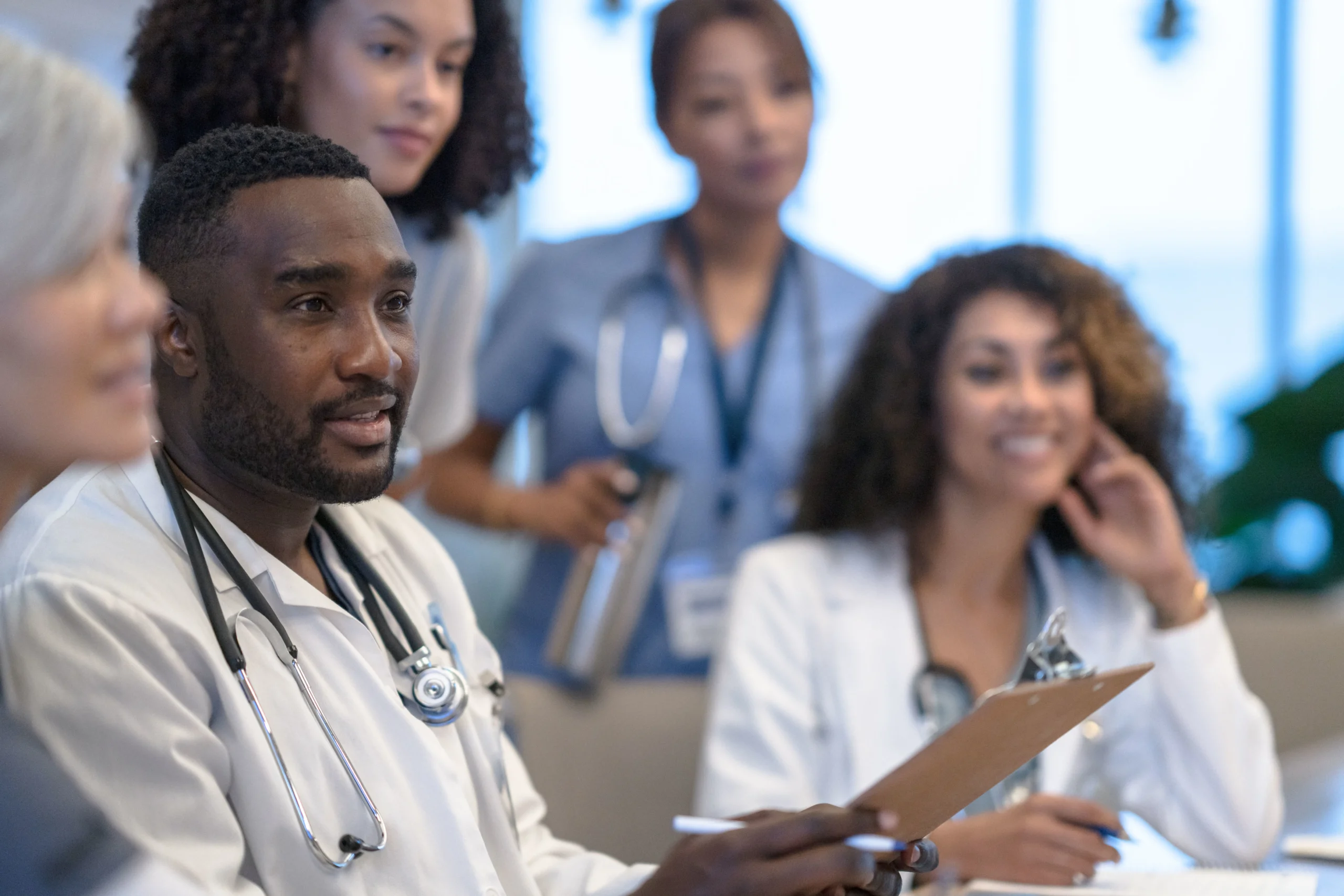 Healthcare team in white coats and scrubs gathers around a table, holding a clipboard and stethoscope, engaged in discussion.