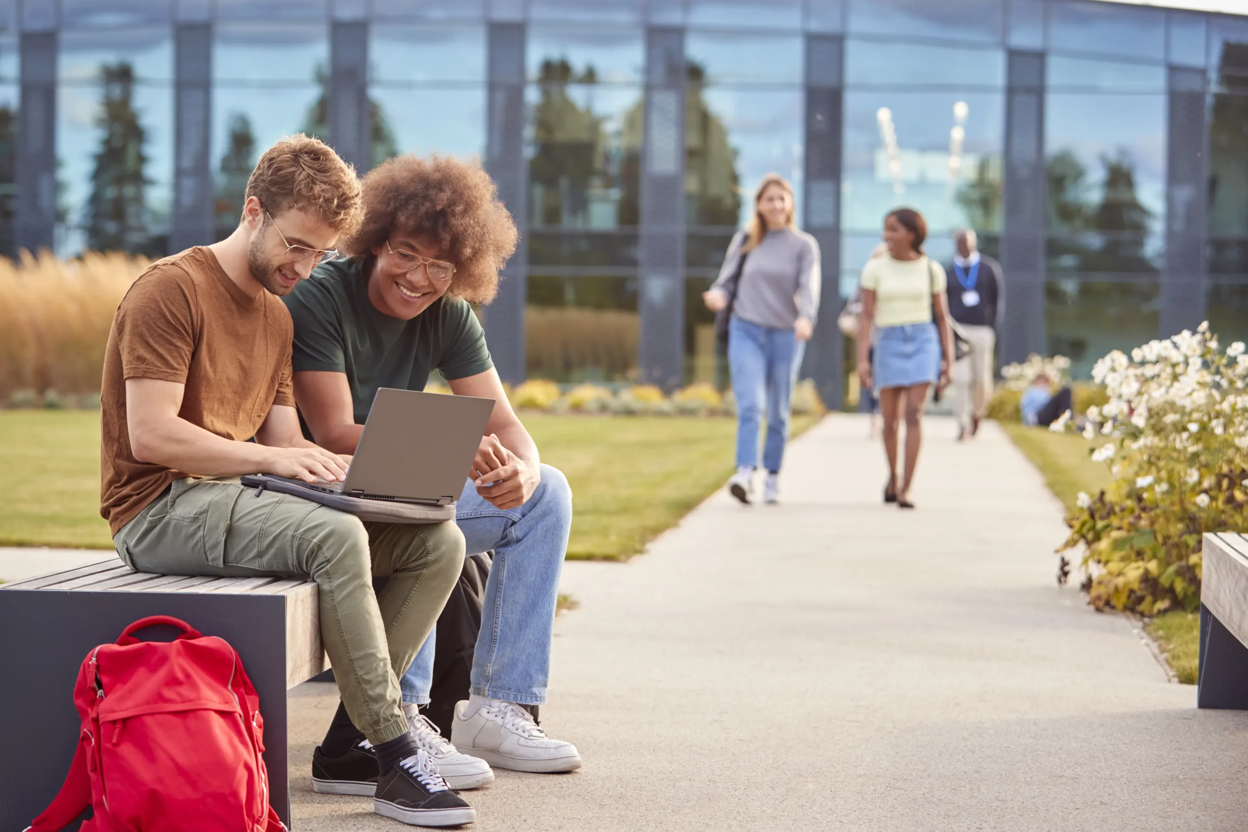 Two young men sit on a bench, smiling at a laptop. One in brown shirt, green pants; other in green shirt, jeans; red backpack nearby.