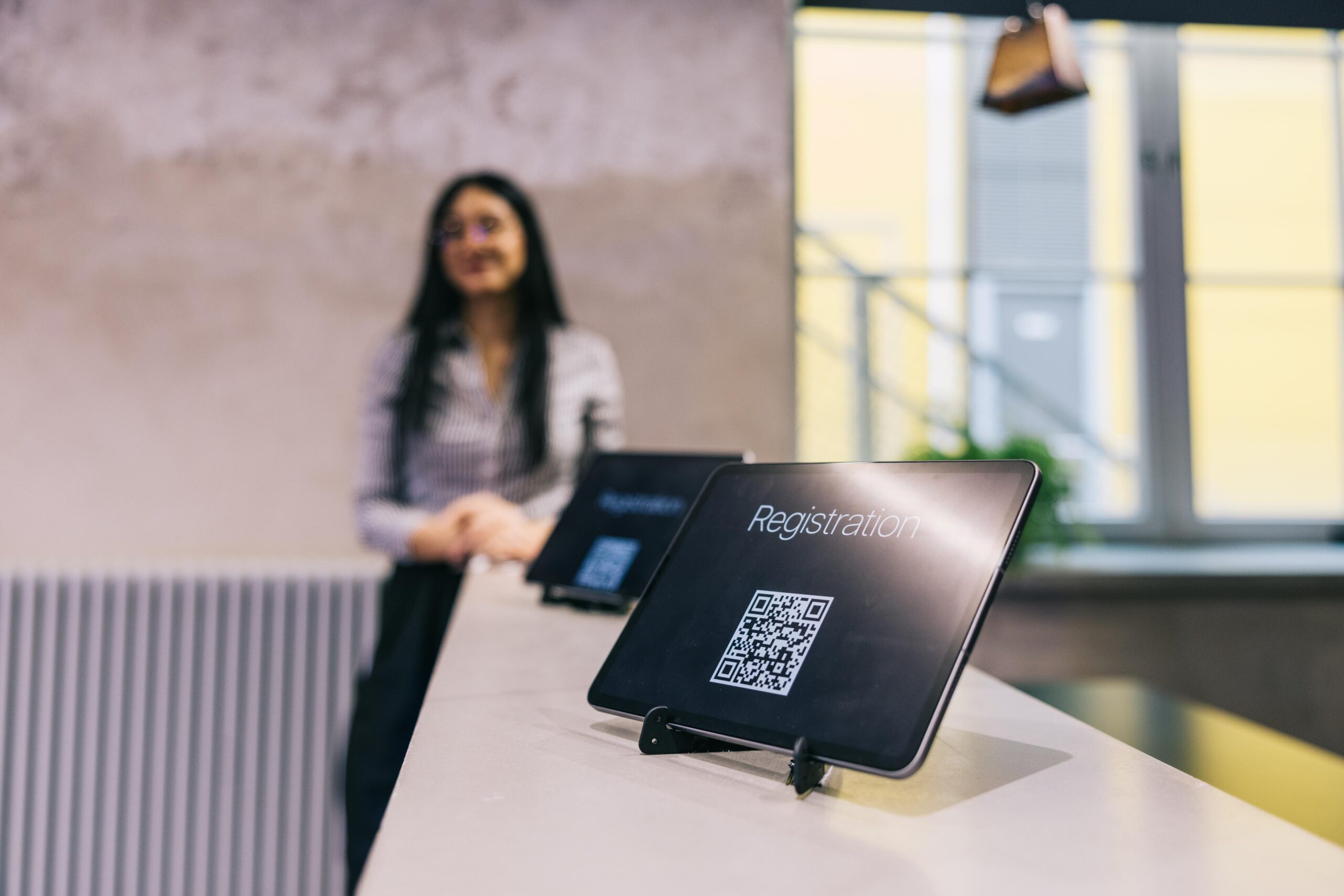 Woman near counter with tablets showing QR codes for event; modern interior, staircase, and large windows in background.