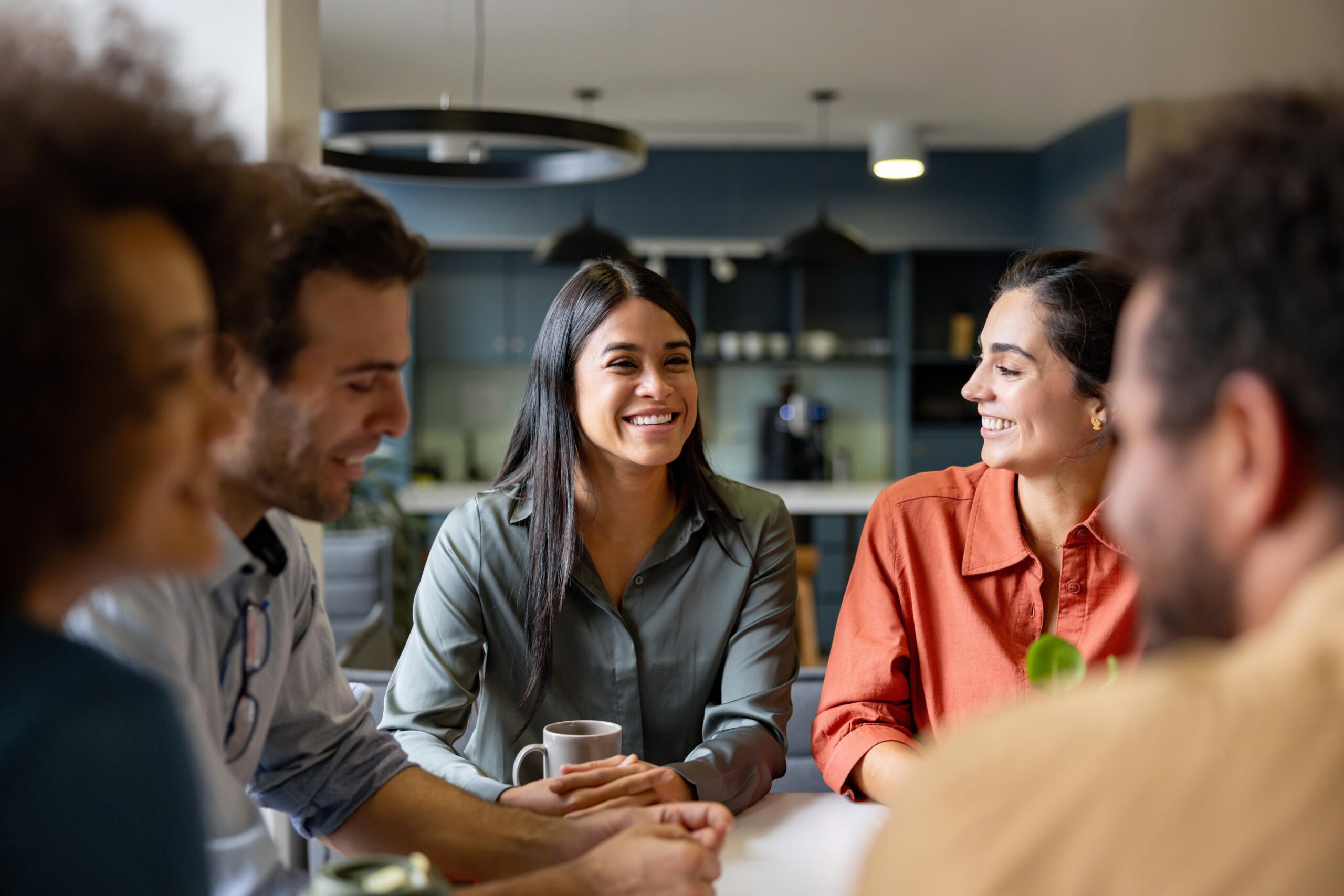 Five people sit around a table, smiling. Focus on woman in gray holding a cup, chatting with woman in orange.