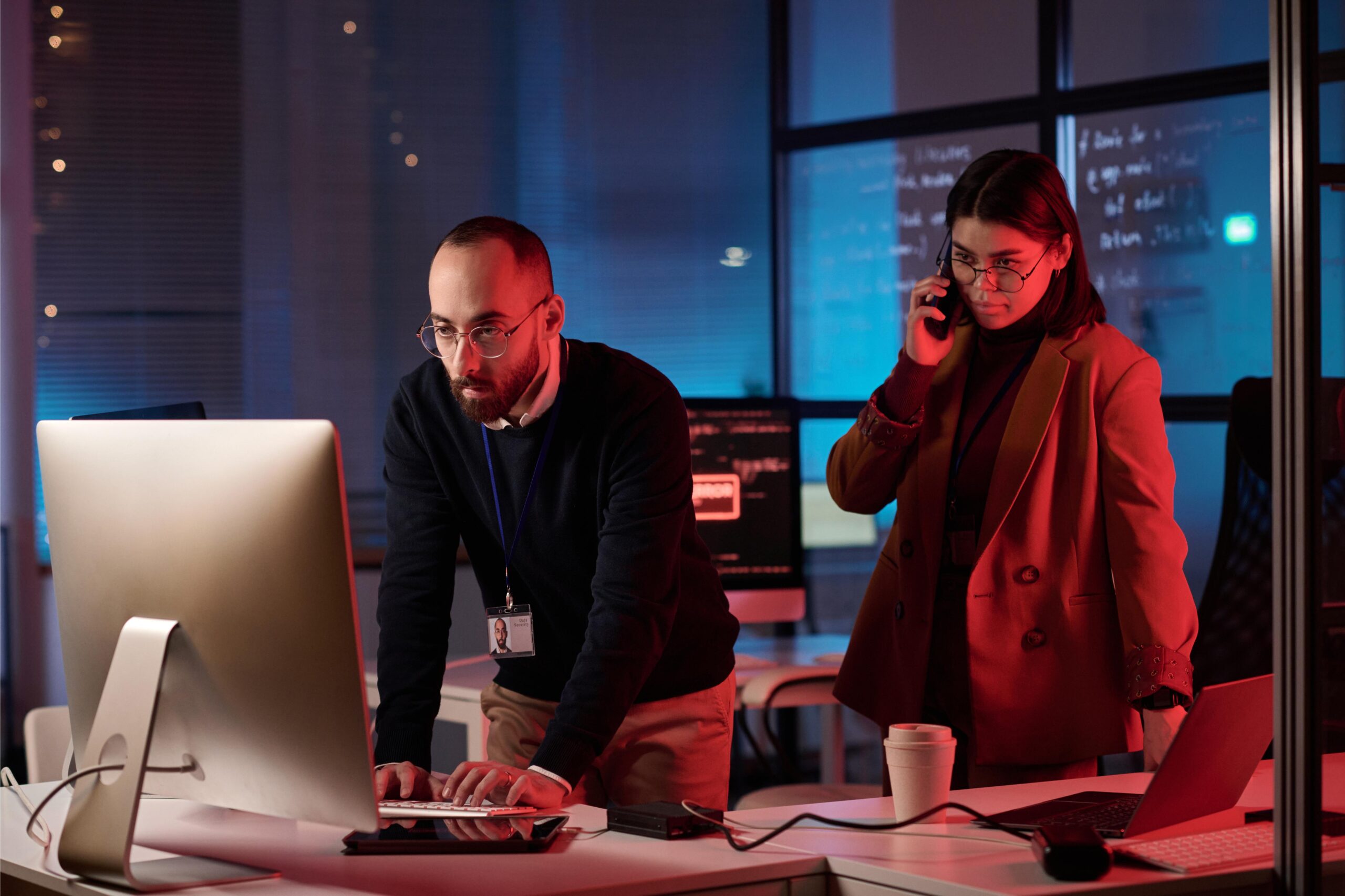 Man types at a computer; woman in brown blazer talks on phone. Dim office, coffee cup on desk.