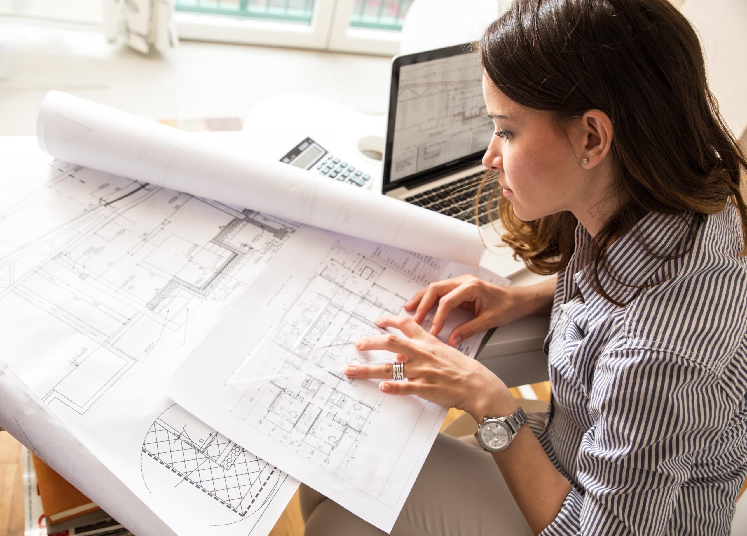 A woman with long brown hair examines blueprints at a desk with a laptop and calculator in a bright room.