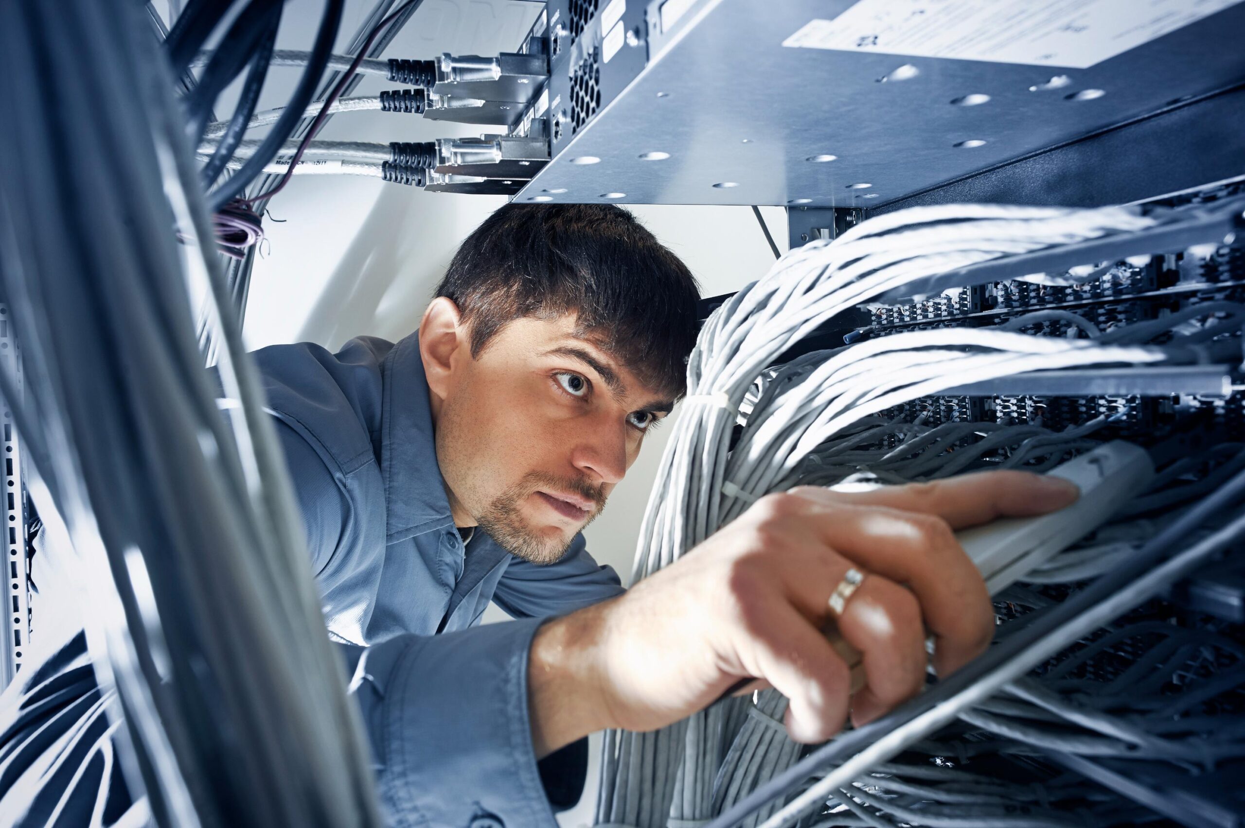 Man in a blue shirt crouched under server rack organizing network cables in dimly lit data center.