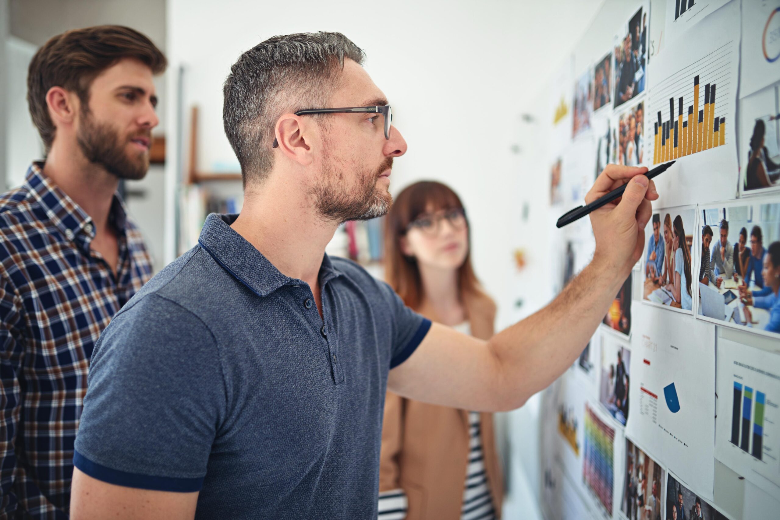 A man with glasses writes on a chart-covered wall, while a woman and another man observe in a collaborative session.