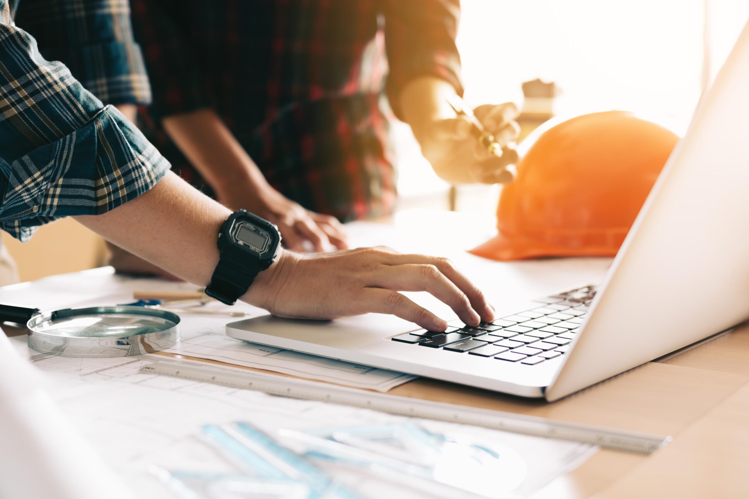 Two people collaborate at a table with a laptop, engineering tools, and an orange hard hat; one types while the other points.