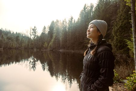 A person in a beanie and puffer jacket looks up by a calm lake with tree reflections; the sky is overcast.