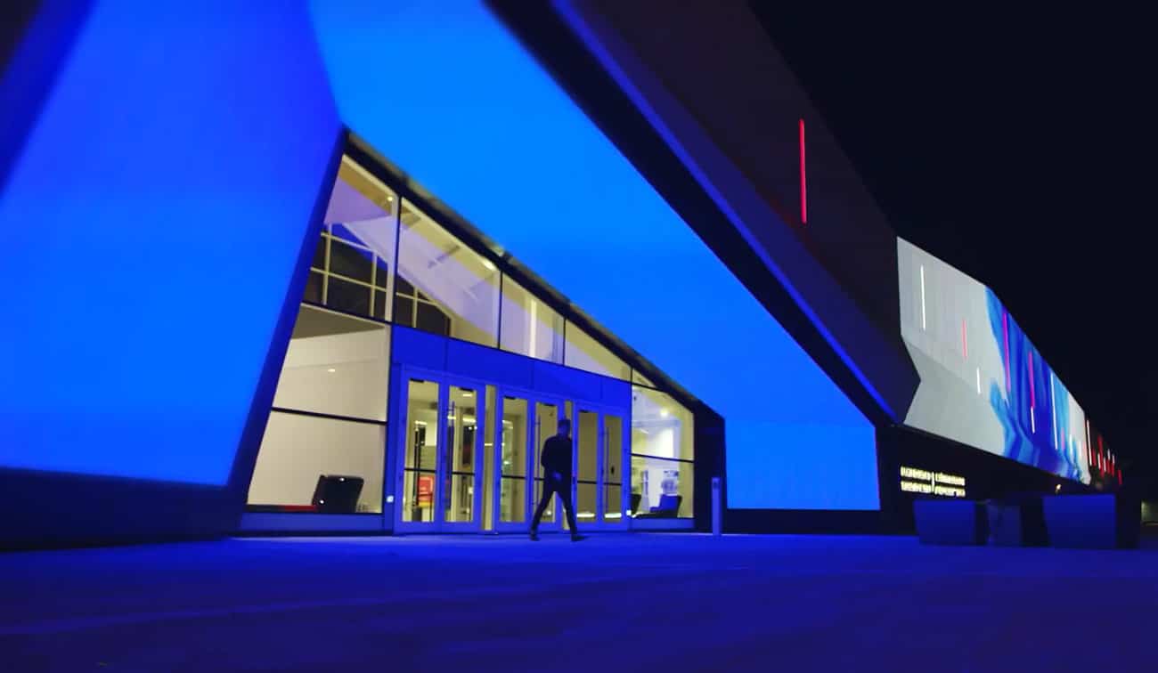 Night view: modern building with striking blue exterior lighting. Person approaches glass entrance; interior brightly lit.