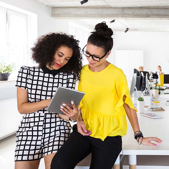 Two women in an office smile while discussing a tablet. One wears black checkered, the other a yellow top; others work nearby.