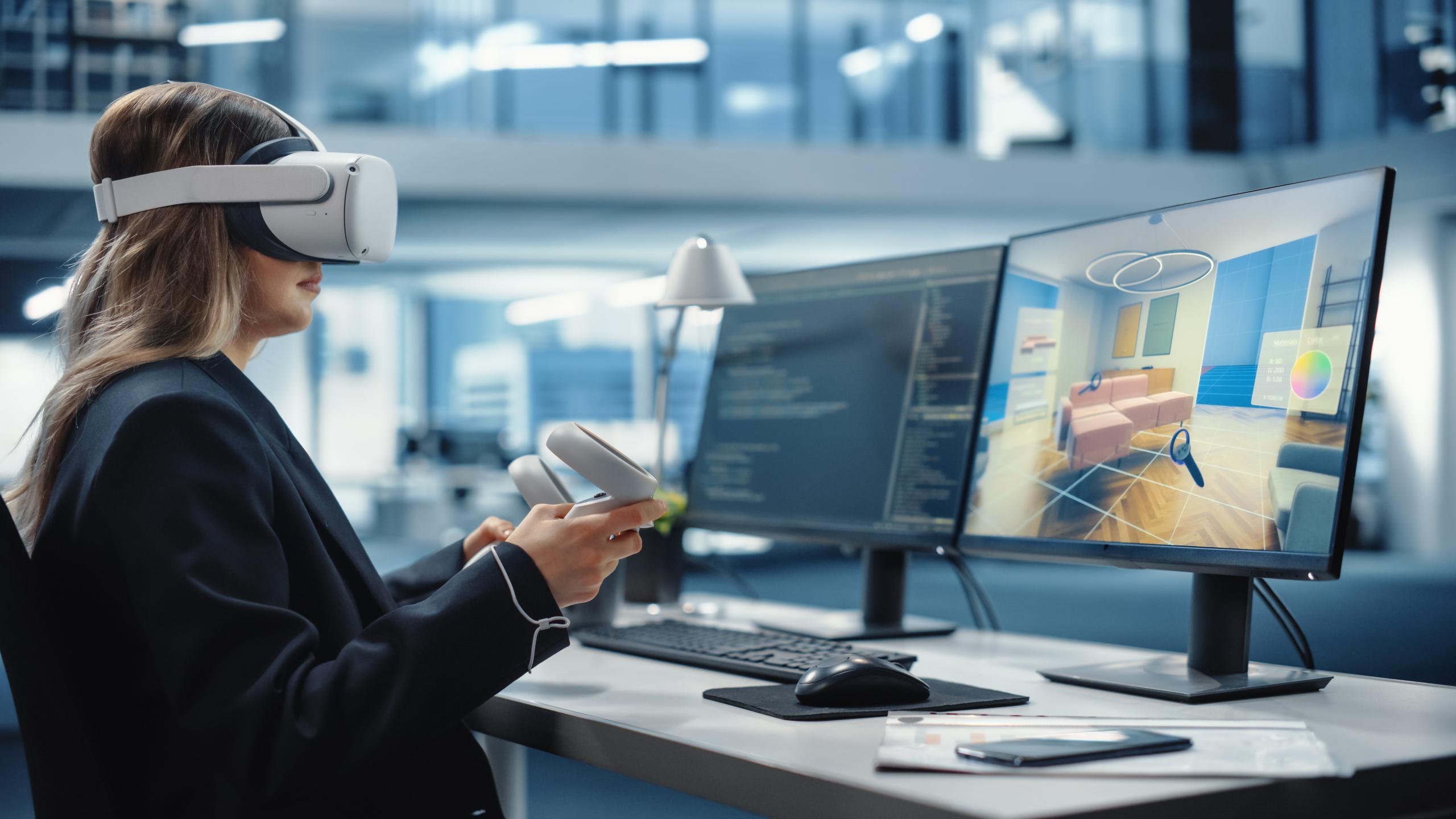 A person with a VR headset sits at a desk, facing monitors showing a 3D room and code; the office background is blurred.