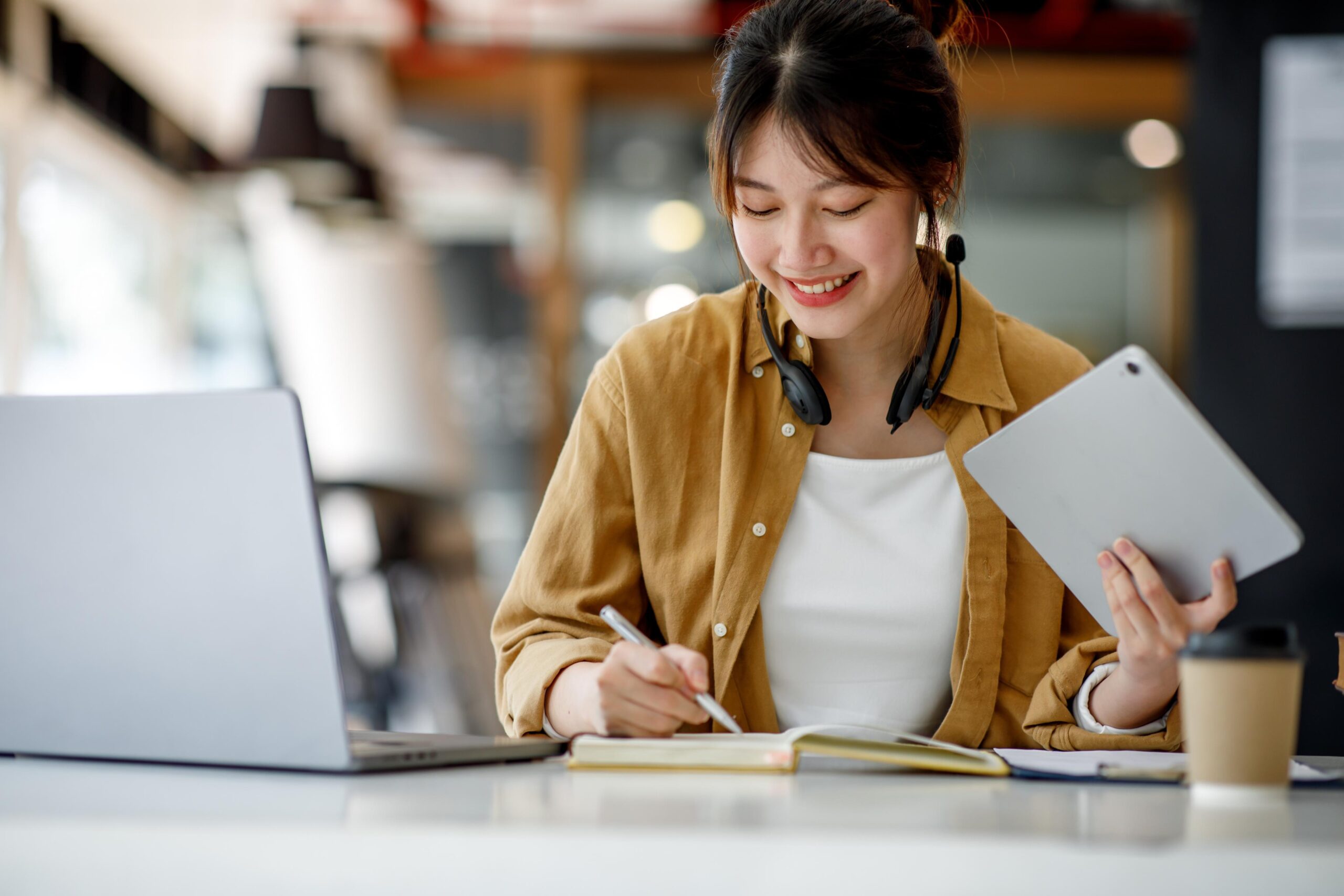 A woman smiles at her desk, writing in a notebook. Tablet in hand, headphones around neck, laptop open with coffee nearby.