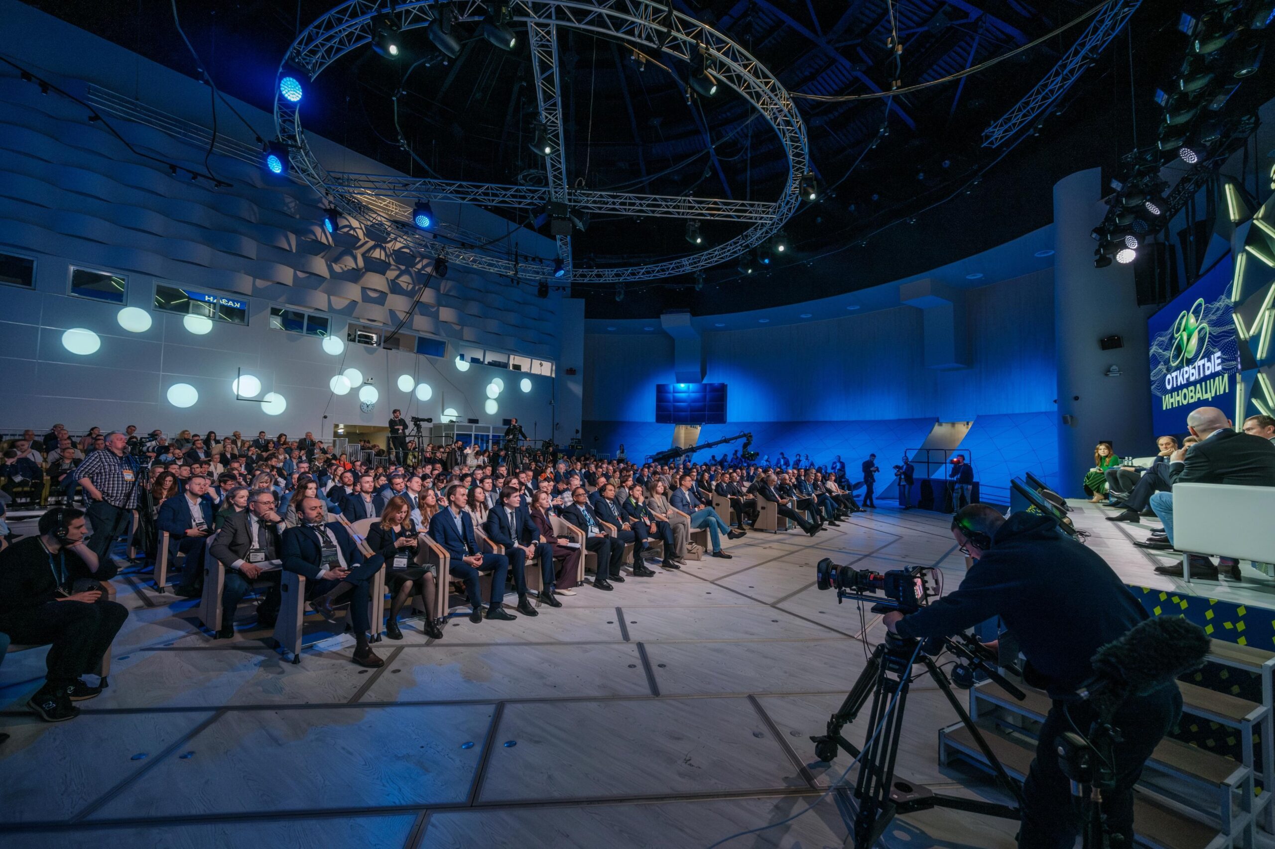 A large audience in blue-lit auditorium watches a panel discussion on stage. Circular lights and complex truss design dazzle.