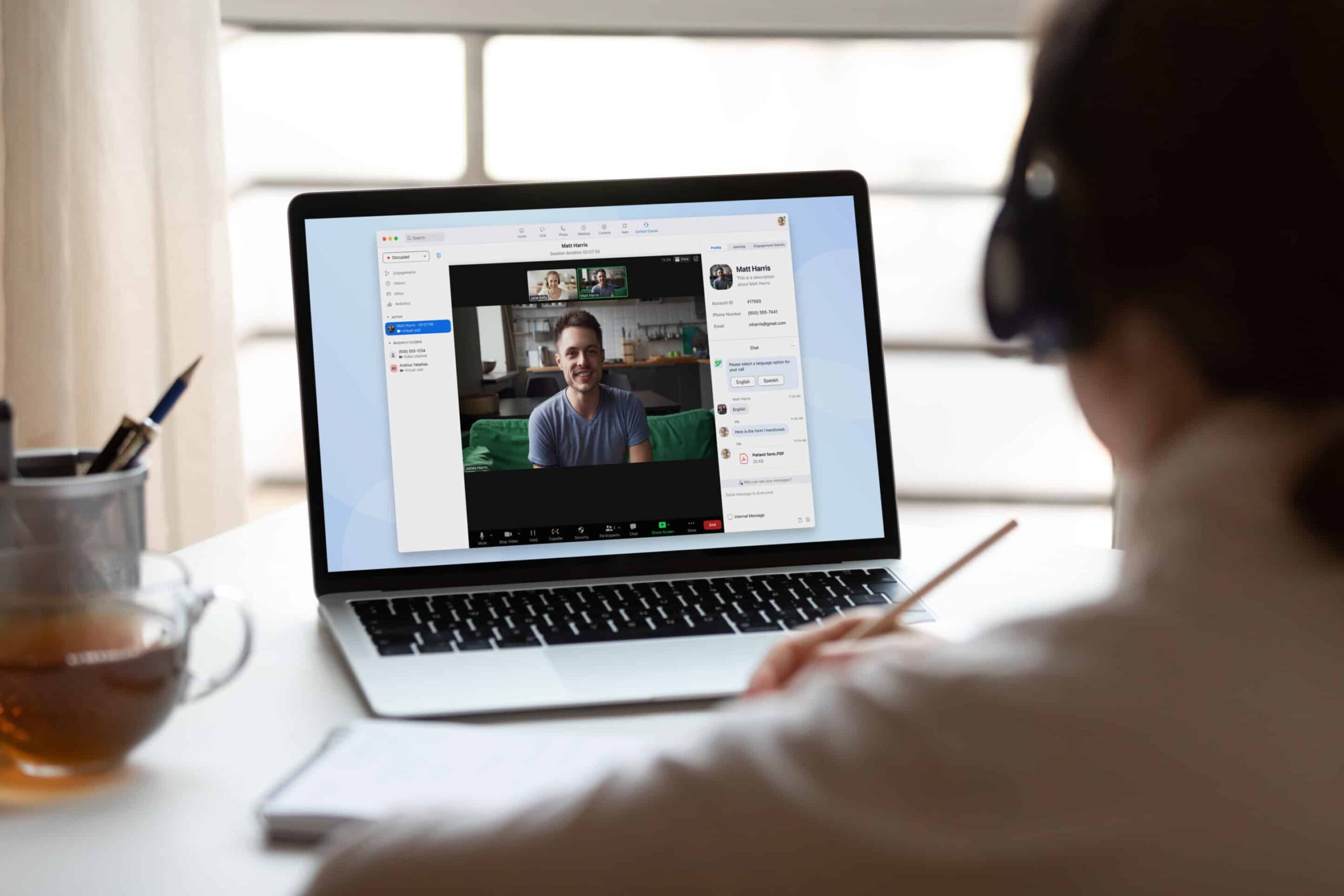 Person on video call with laptop, headphones on. Screen shows another person. Table has tea and a notebook.