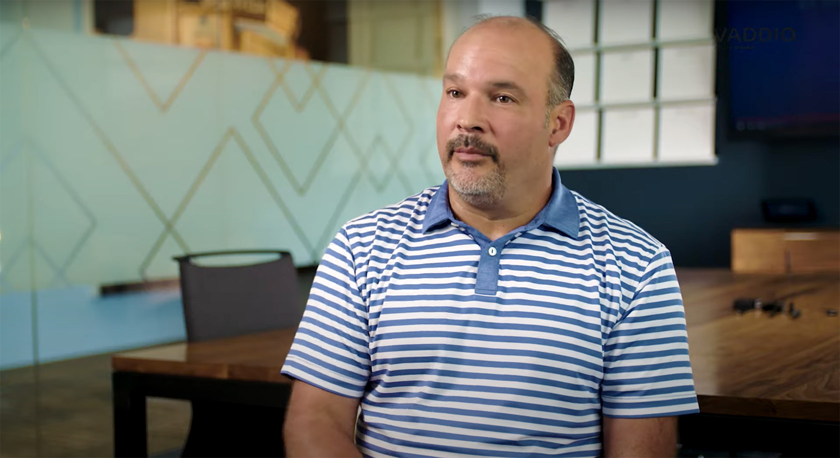 A man wth striped t-shirt sat in front of camera talking.