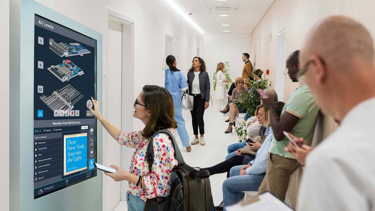 People wait in a hallway; some seated, others on devices. A woman checks a 22Miles digital directory, phone in hand. Office-like setting.