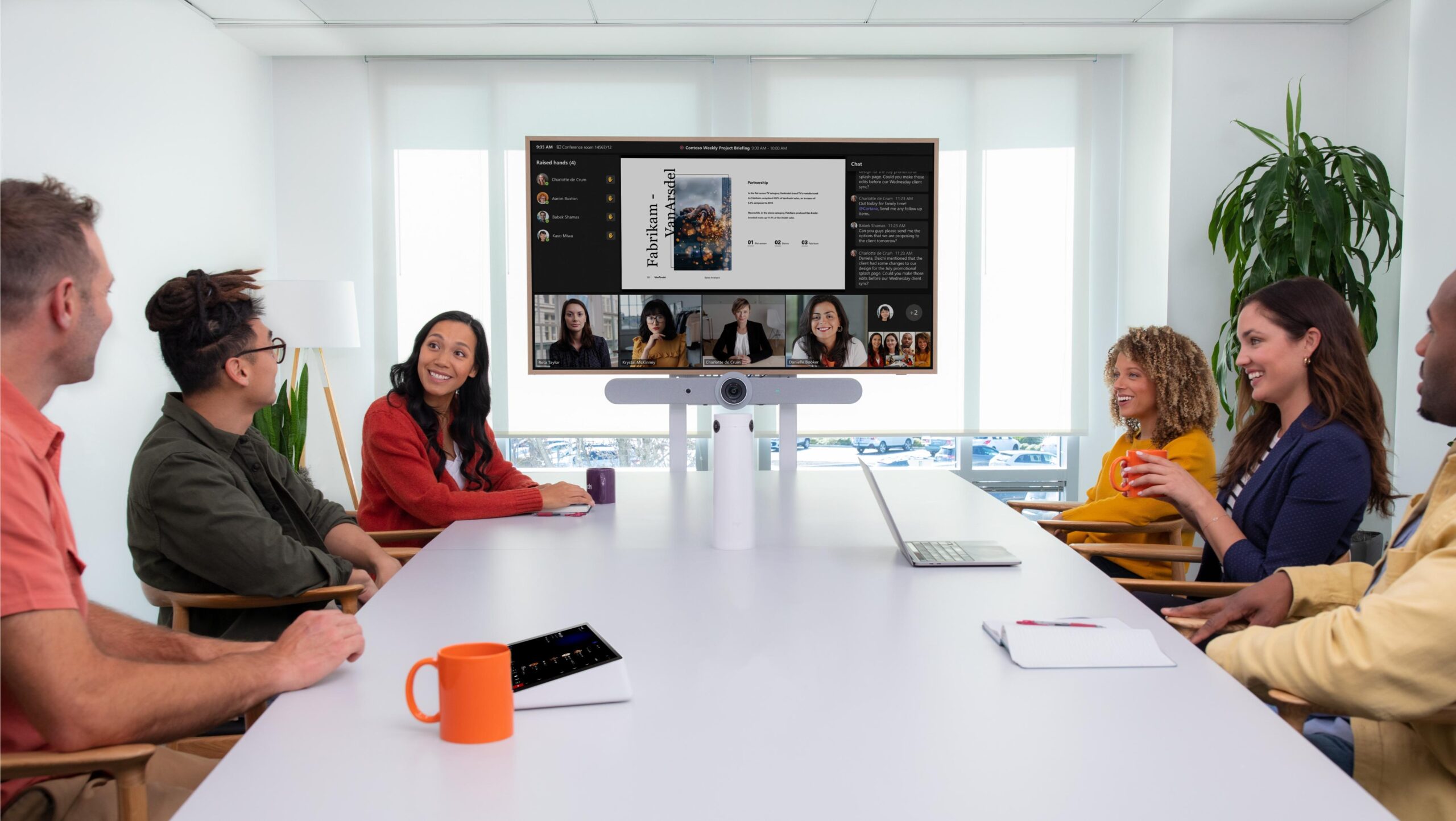 Six diverse people sit around conference room with a video conference on the screen; a Logitech bar, laptops and mugs scattered about.