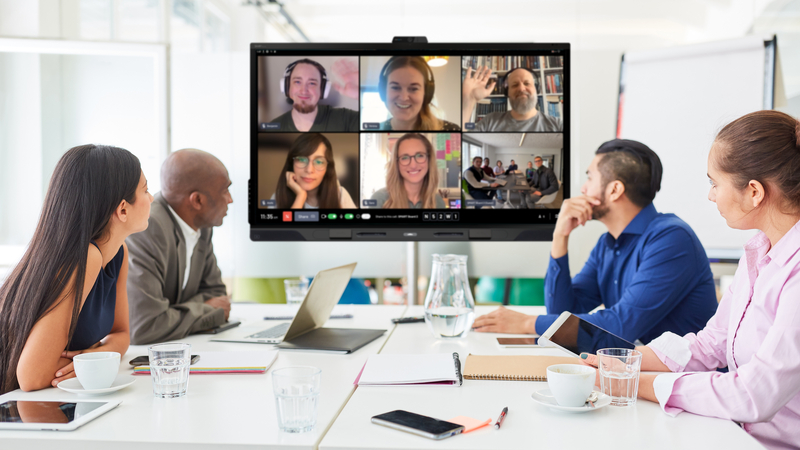 Four people in a conference room use a SMART board to host a hybrid meeting with remote attendees on screen.