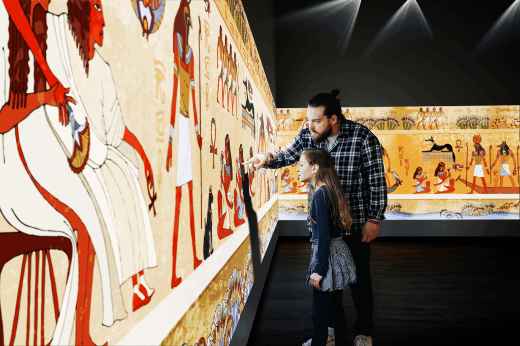 A father and his daughter look at and touch an immersive projection of Egyptian art.